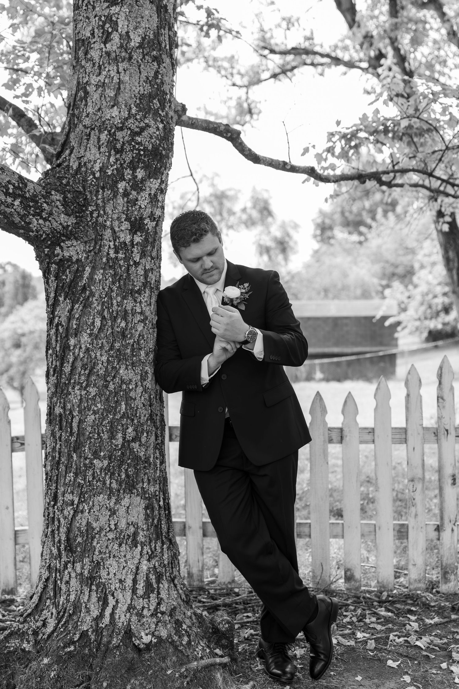 Groomsman in black suit poses playfully behind tree trunk in outdoor wedding setting.