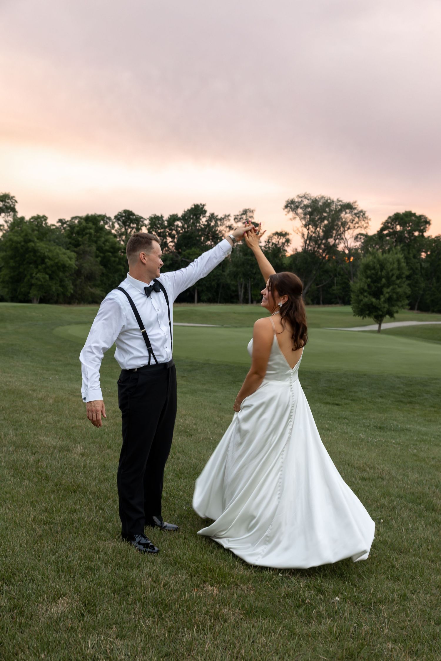 Bride and groom dancing outdoors on green grass at sunset, groom in suspenders spinning bride in white gown.