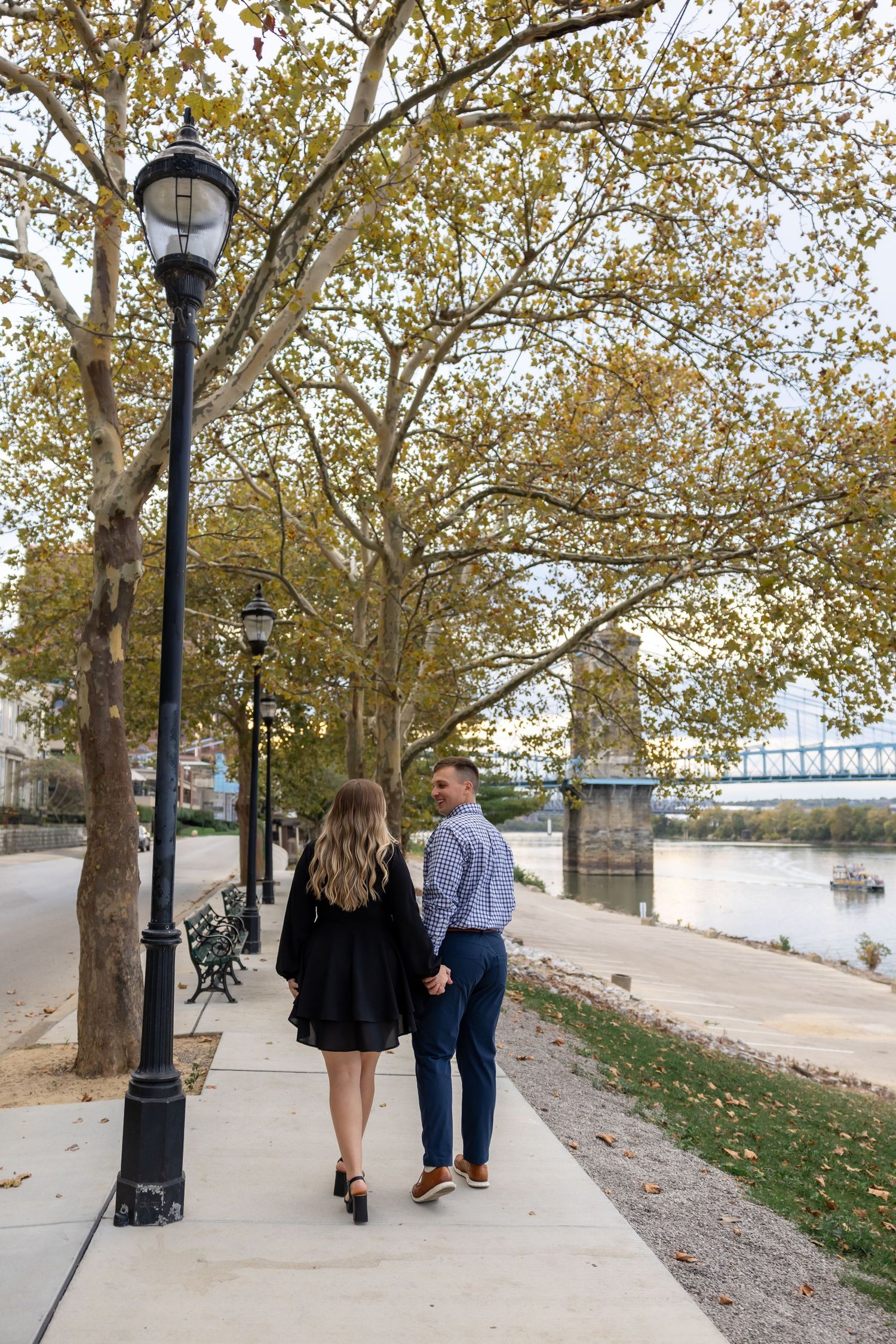 Couple walks along a tree-lined riverside path with lampposts at sunset.
