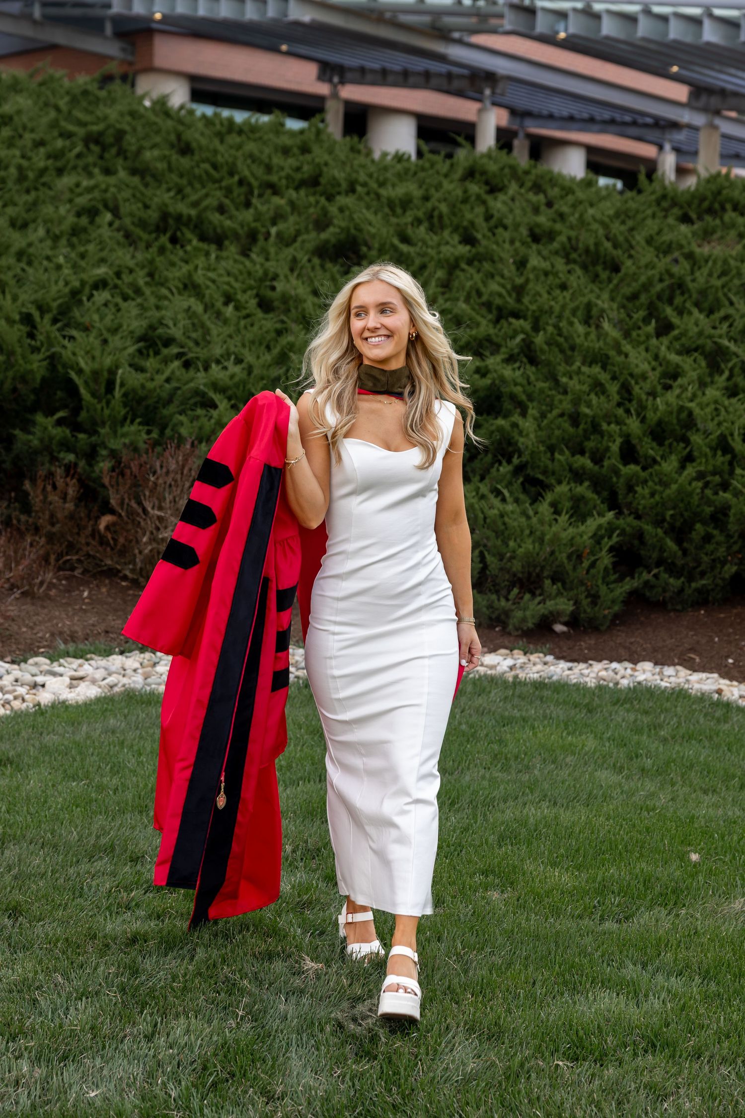 Young woman in white dress holding red doctoral graduation gown, walking on green lawn near evergreen shrubs.