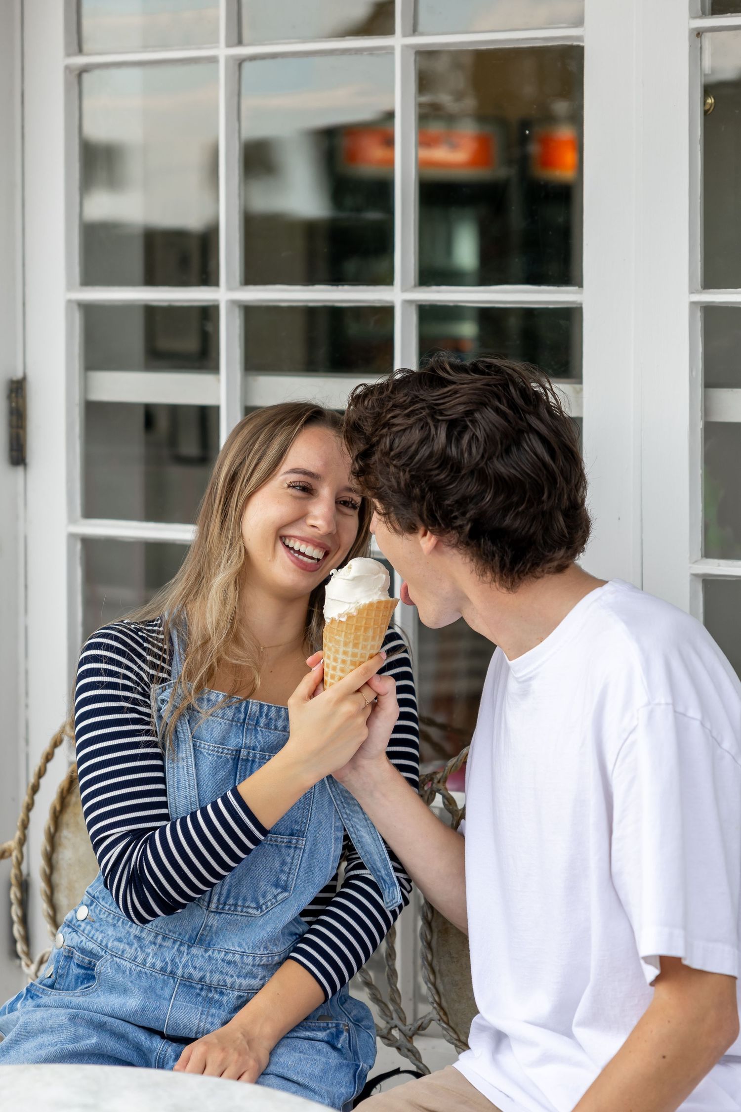 Couple sharing ice cream and laughter on a porch with white windows.