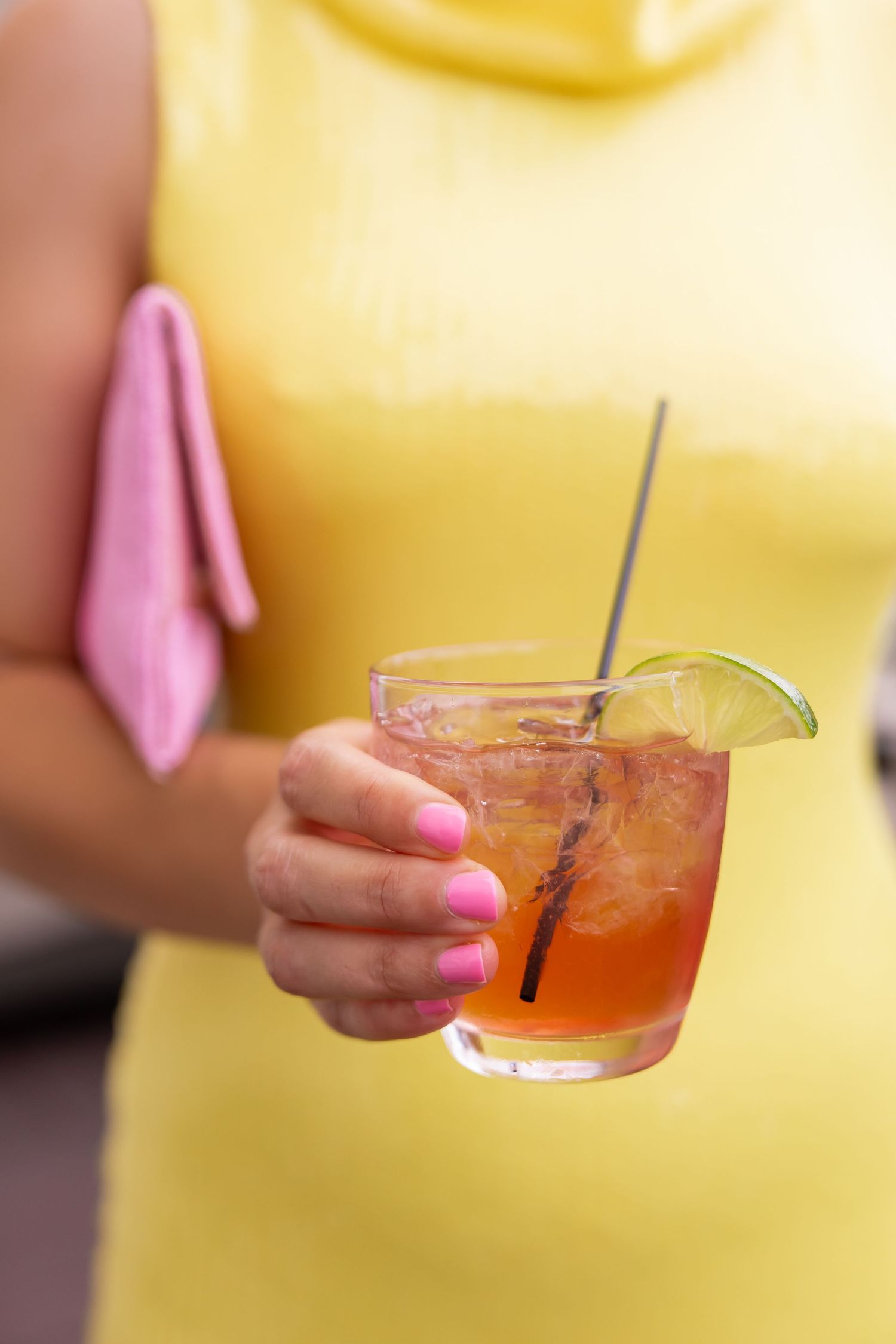 Woman in yellow dress holding a pink cocktail with lime garnish and straw.