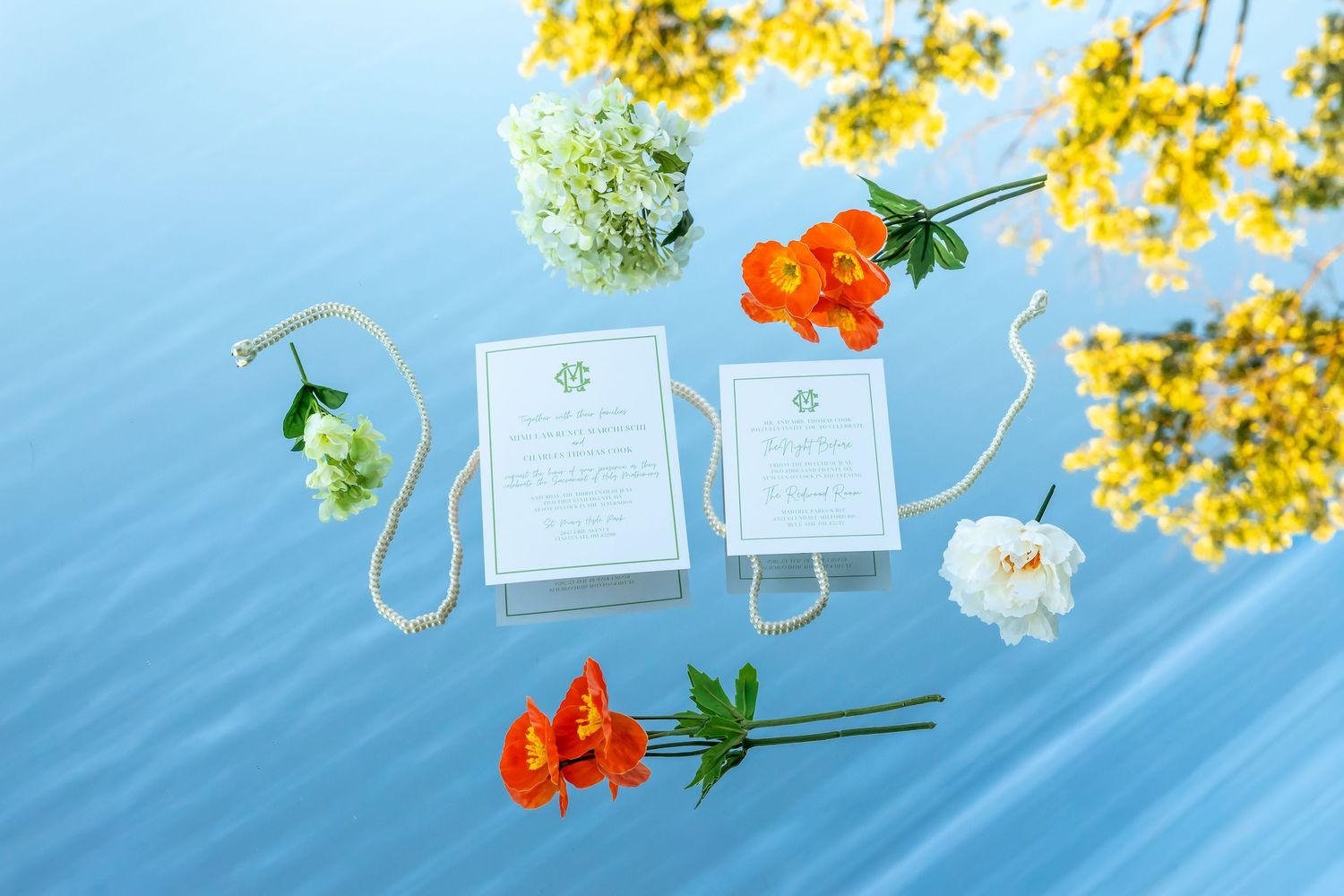 Elegant wedding stationery flat lay with orange poppies, white peonies, and yellow flowers on blue water.