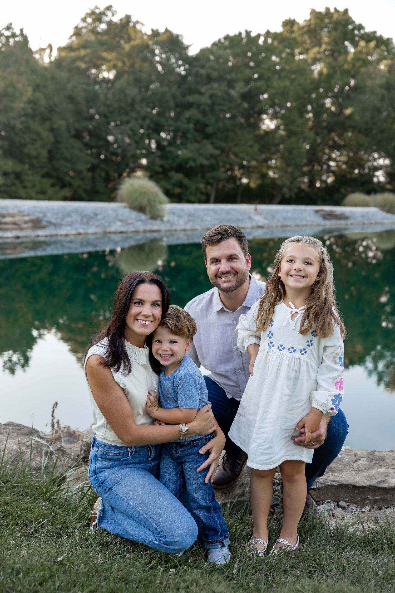 Family relaxes together on grassy area near a serene lake at sunset.