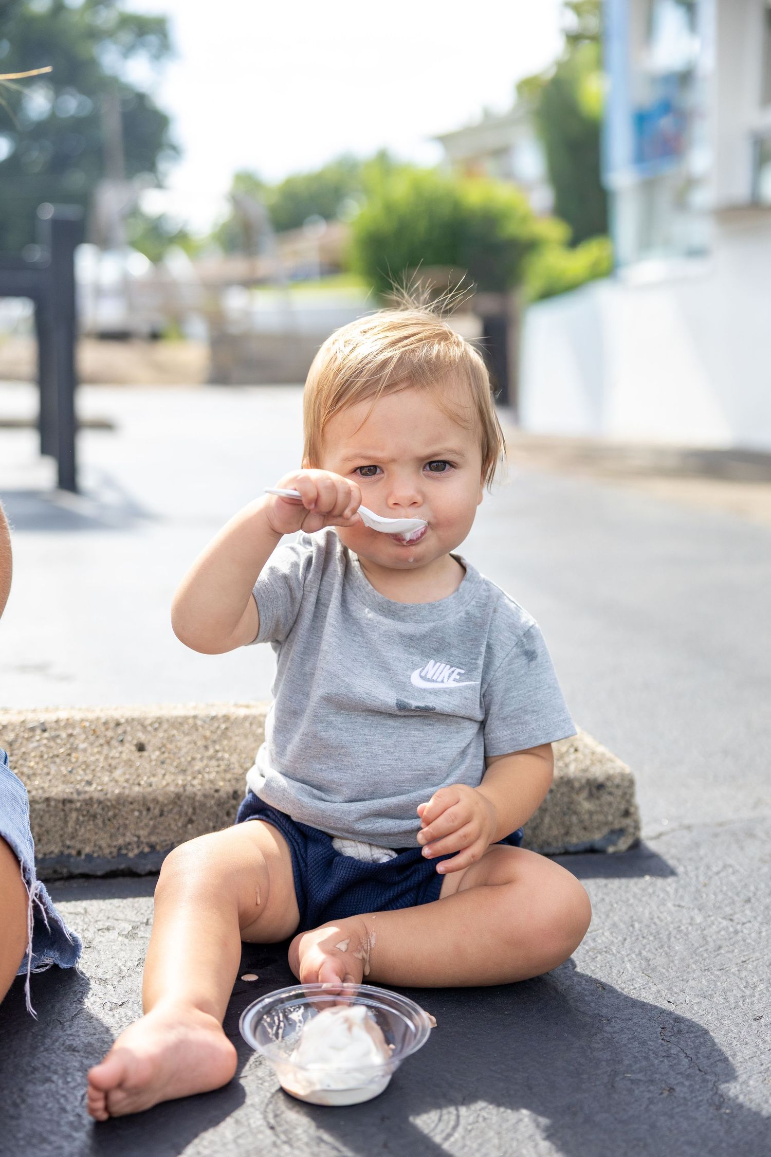 Small child in grey shirt sits on concrete surface eating a snack.