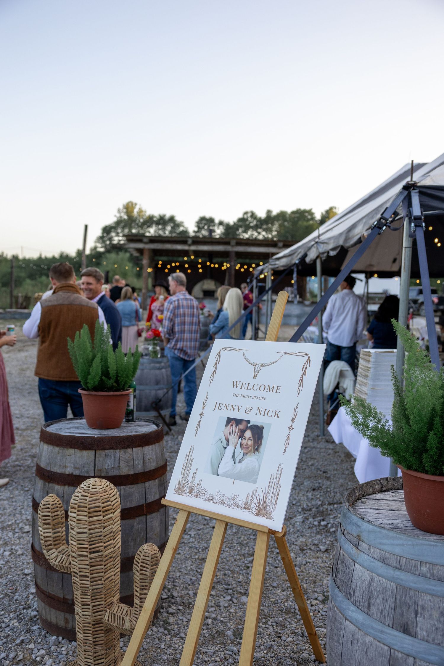 Rustic outdoor wedding reception setup with wooden barrels and welcome signs at sunset.