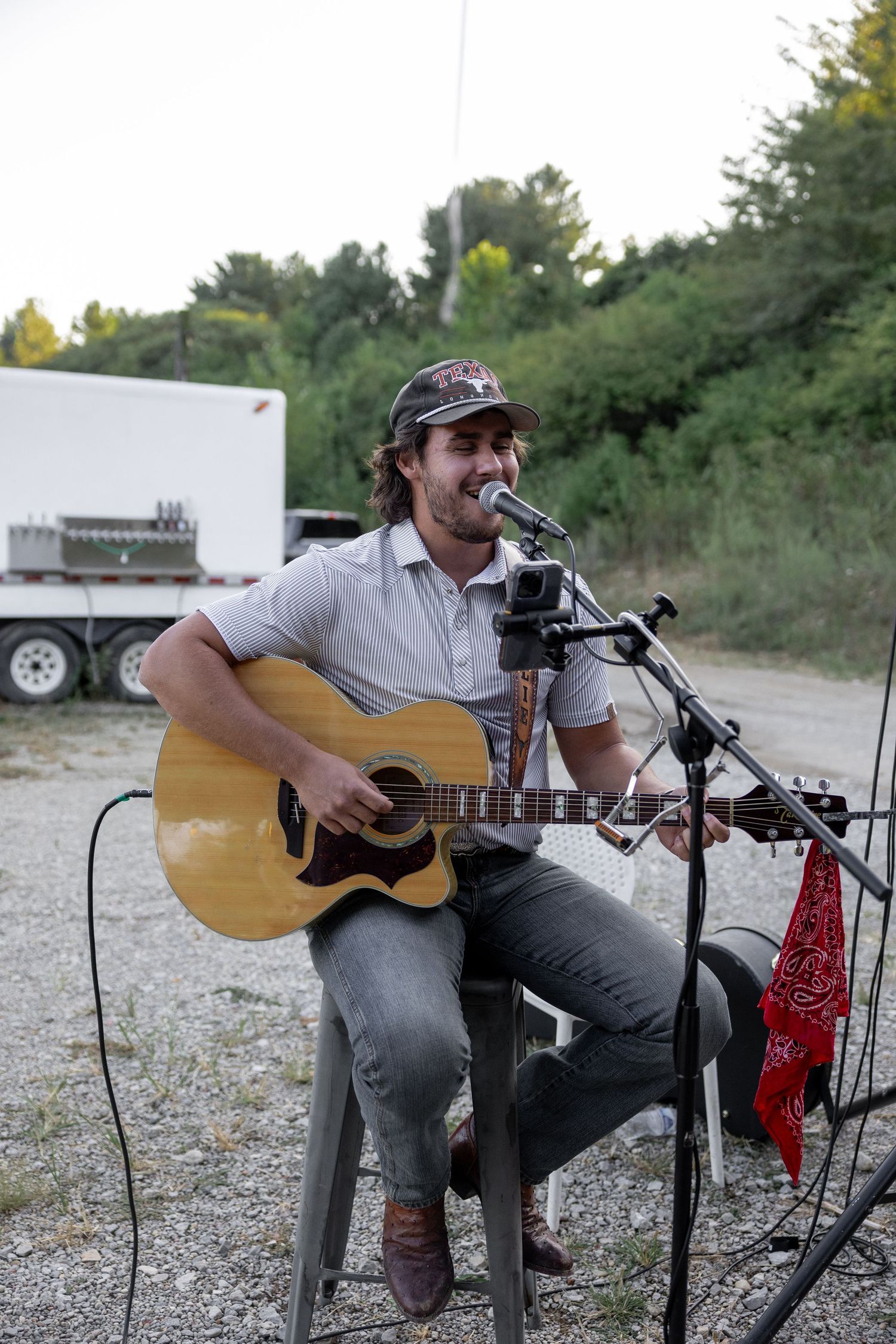 Musician playing acoustic guitar while seated outdoors with microphone setup.