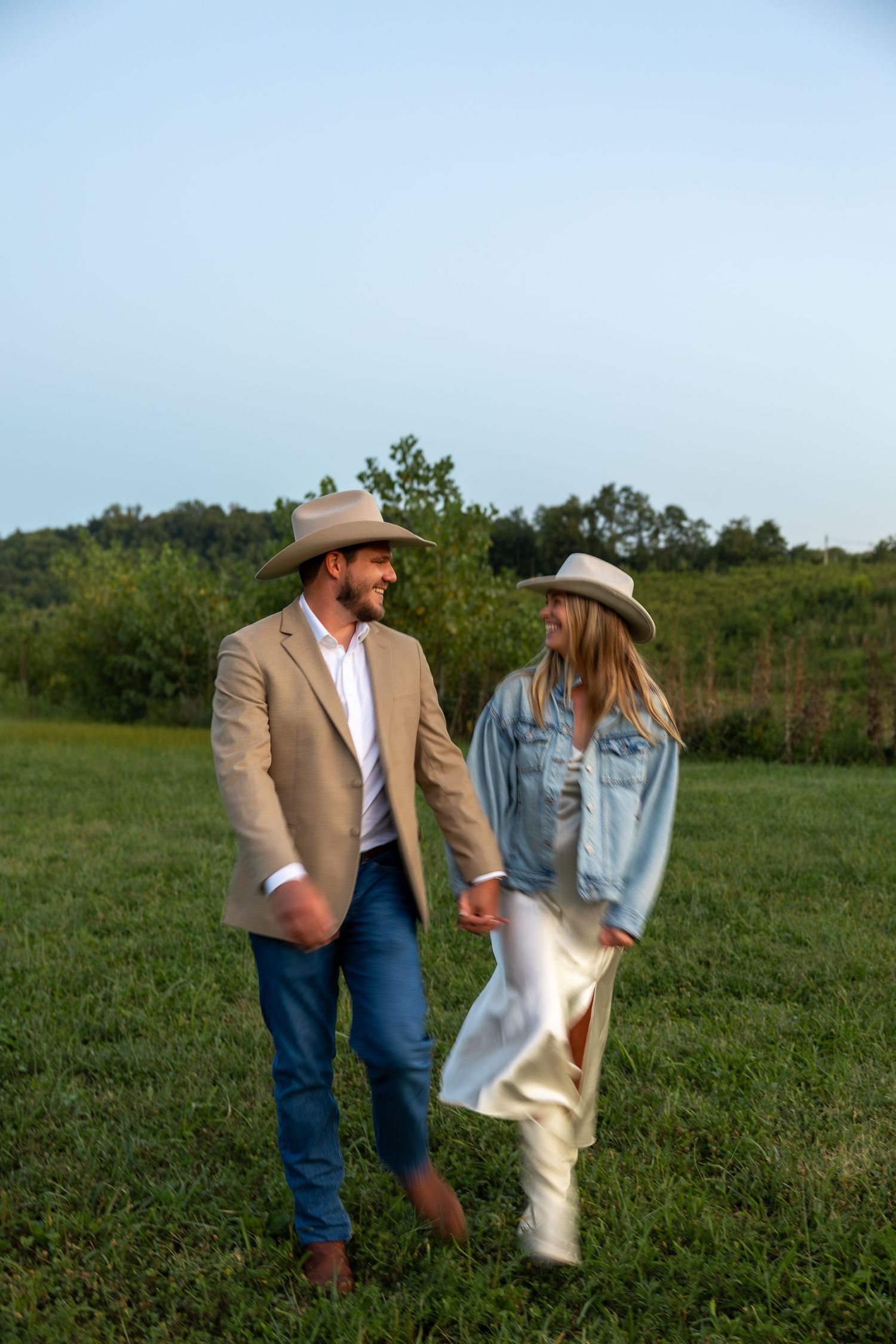 A couple walks through a grassy field at sunset wearing country western attire.