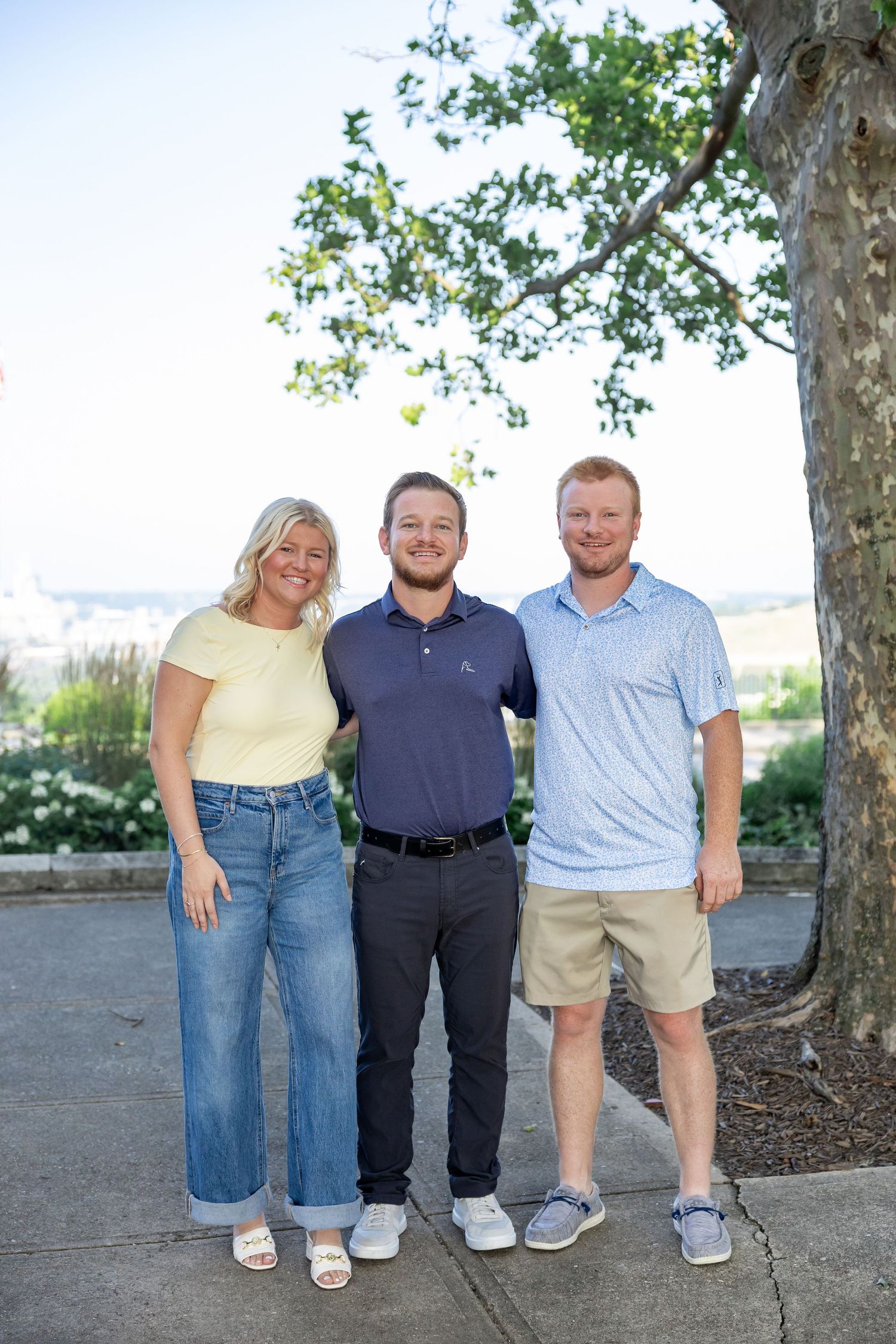 Group photo of three people standing together under trees with water view in background.