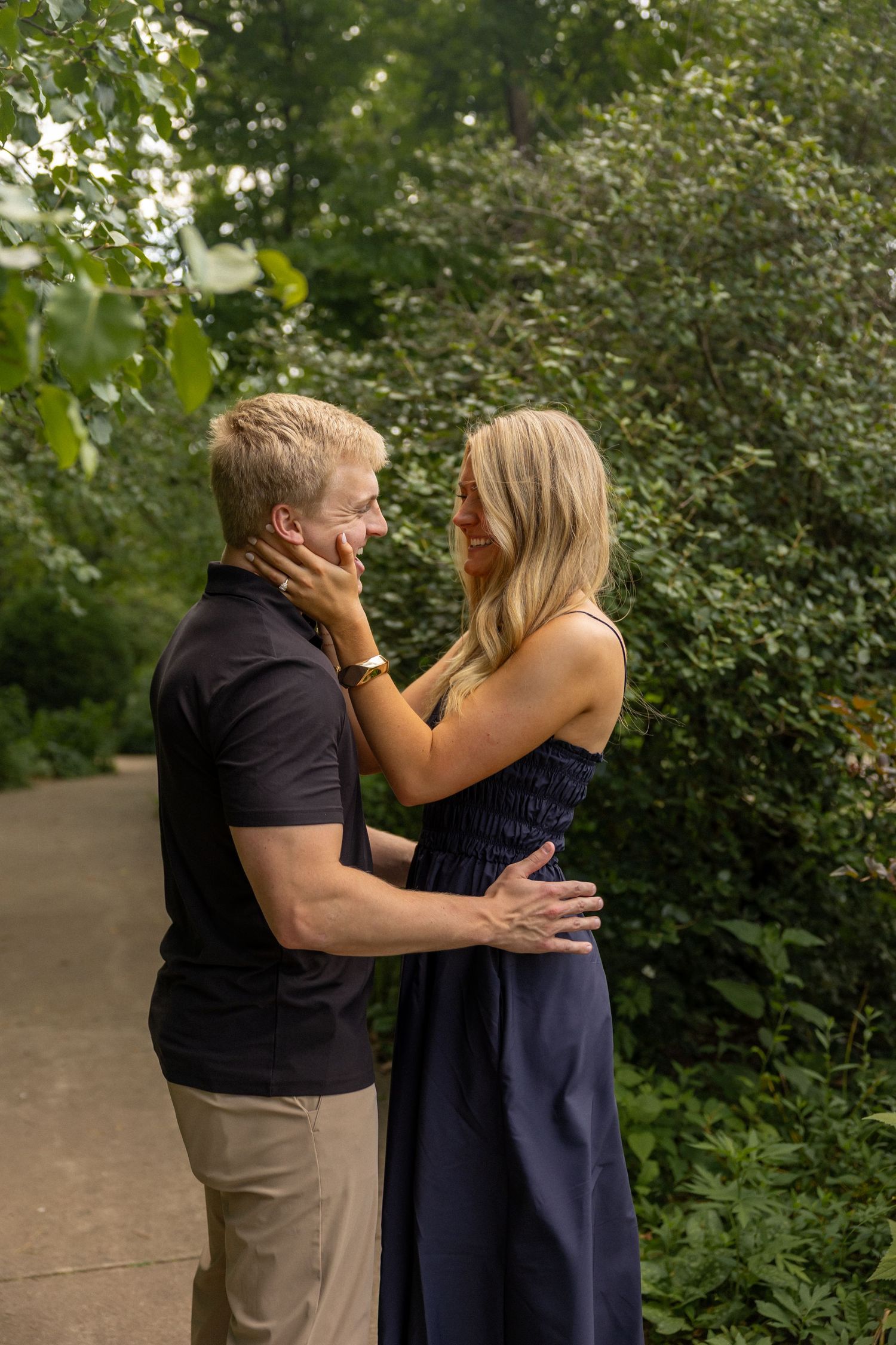 A couple embraces outdoors, woman in navy dress touching man's face, surrounded by lush green trees.