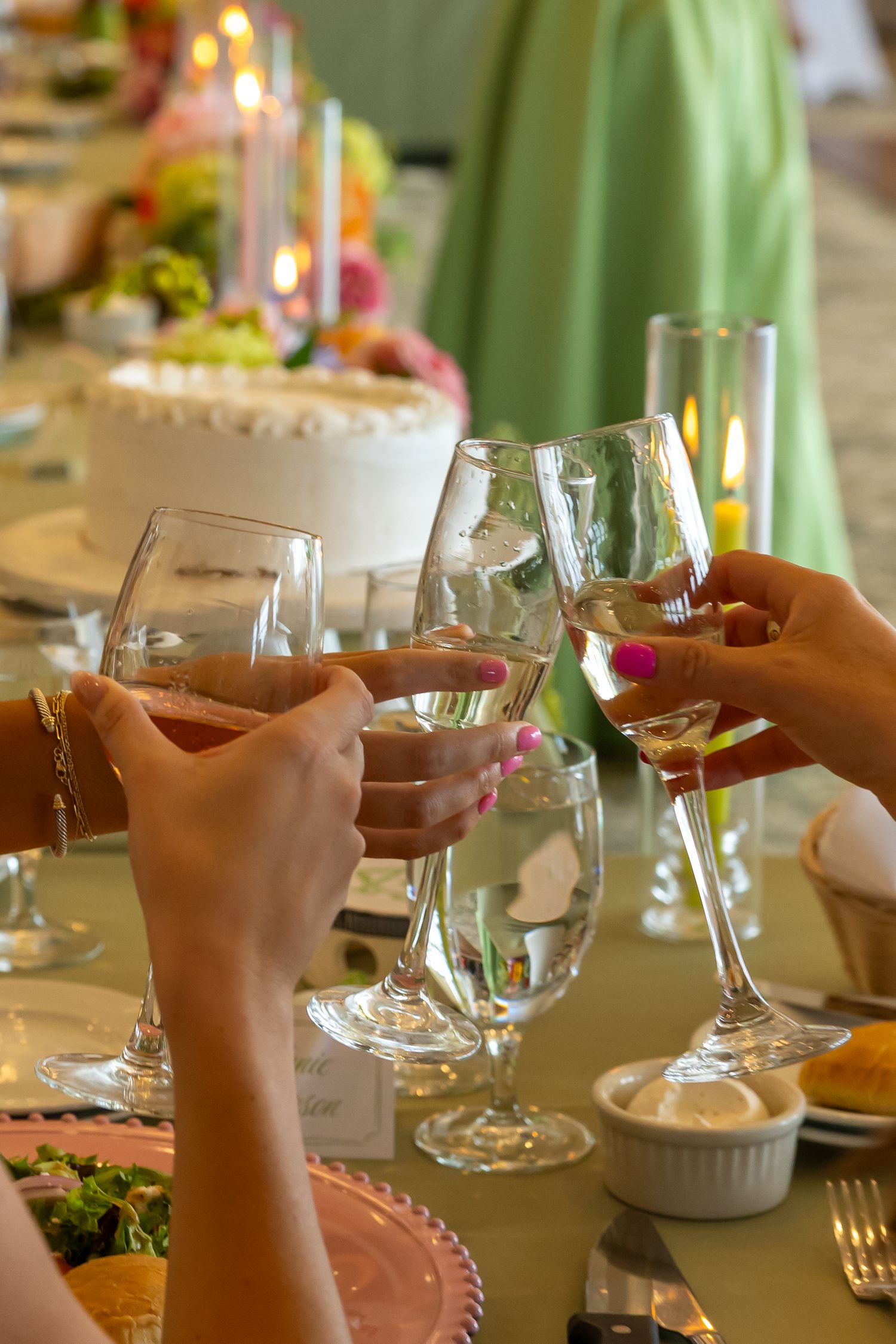 Women clinking wine glasses at an elegant decorated table with candles, a white cake, and pink place settings.