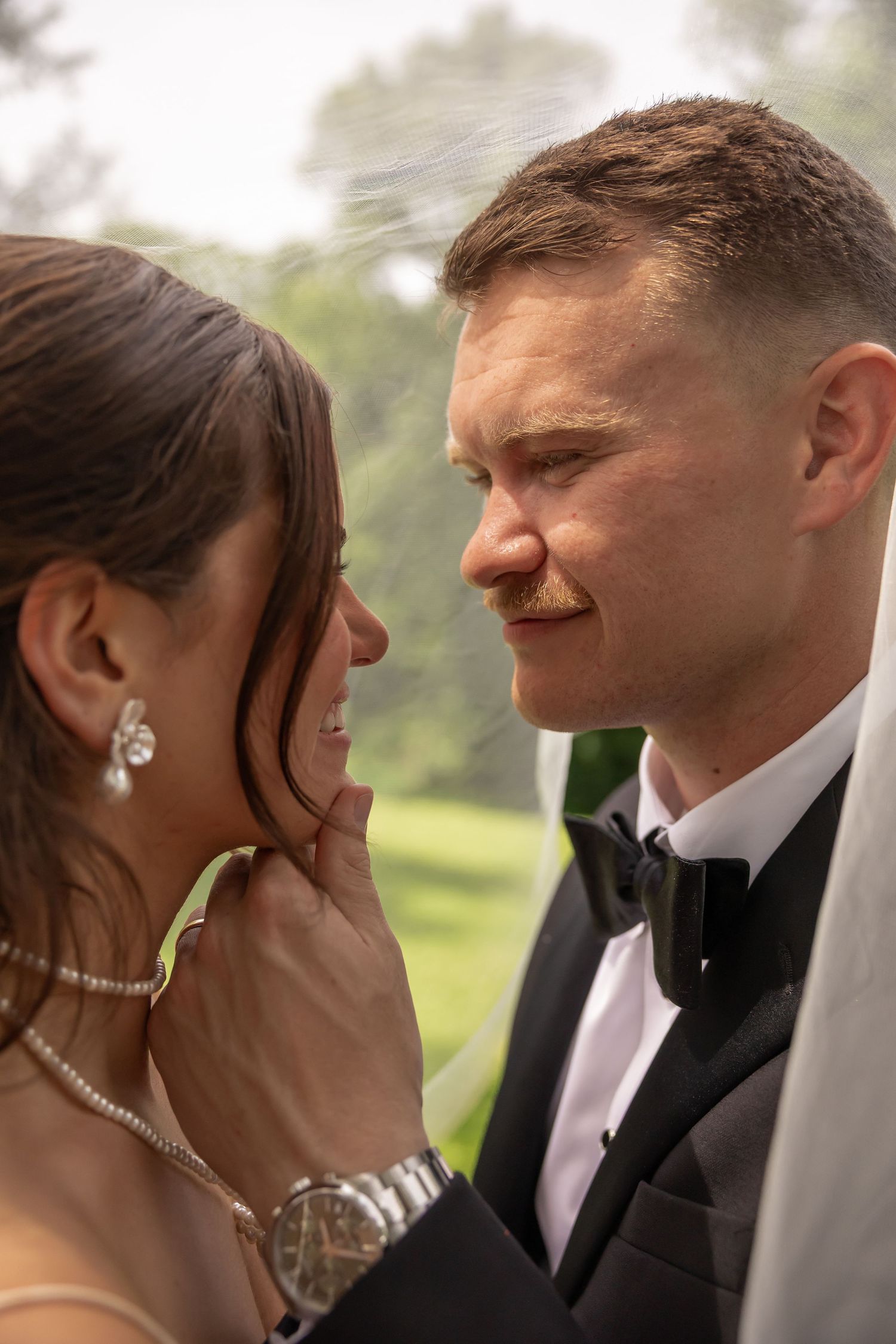 Groom in black tuxedo gazes lovingly at bride wearing diamond jewelry on a misty outdoor wedding day.