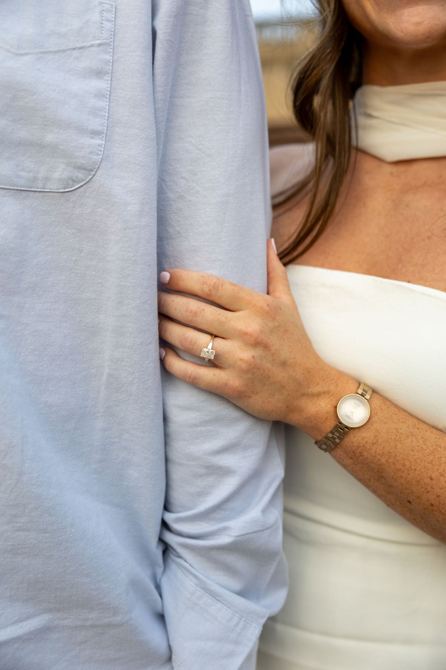 Close-up details of a light blue shirt sleeve and white top with an engagement ring visible.