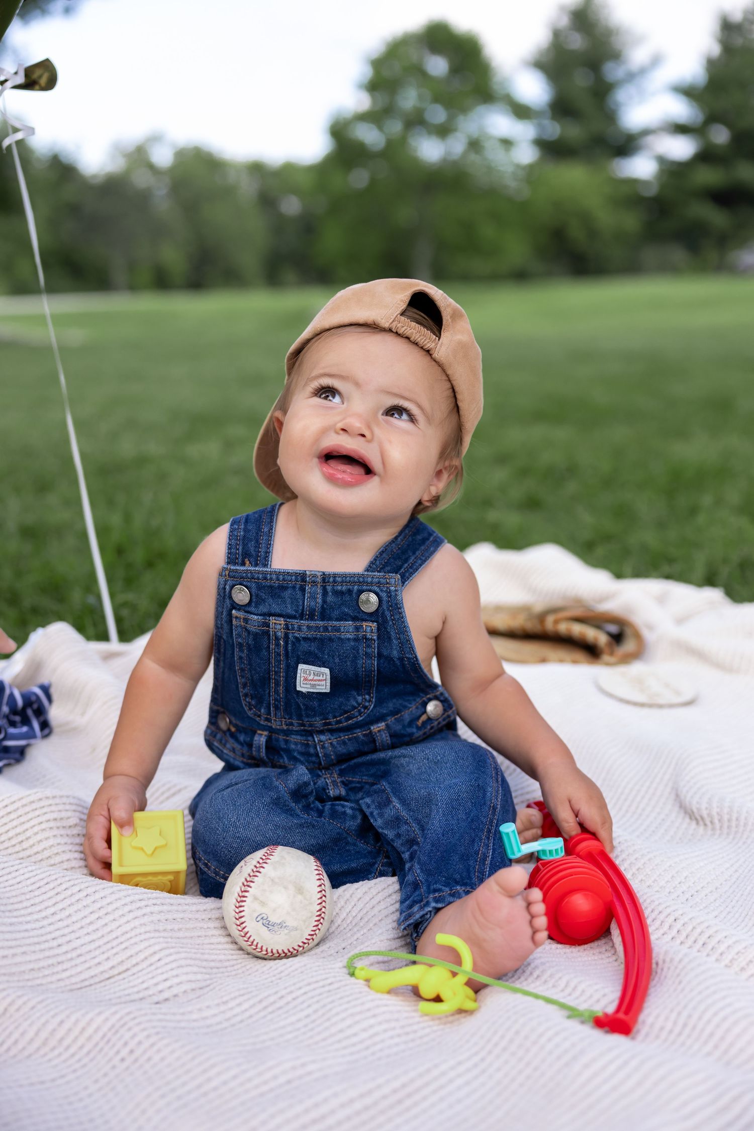 Baby in denim overalls playing with colorful toys on a white blanket outdoors.