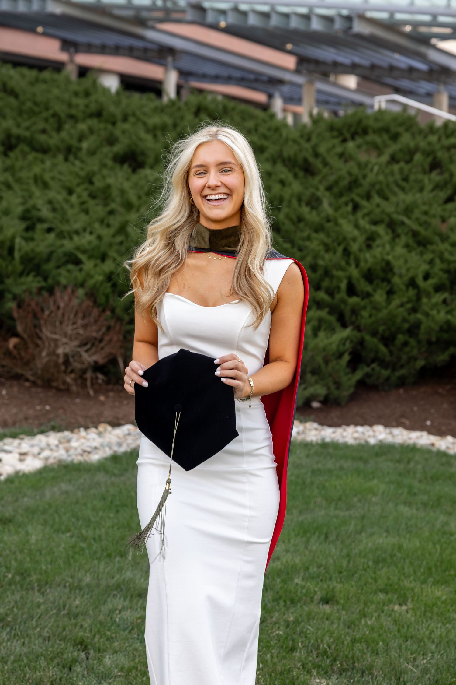 Smiling blonde graduate in white dress holding cap with red hood outdoors near green bushes.