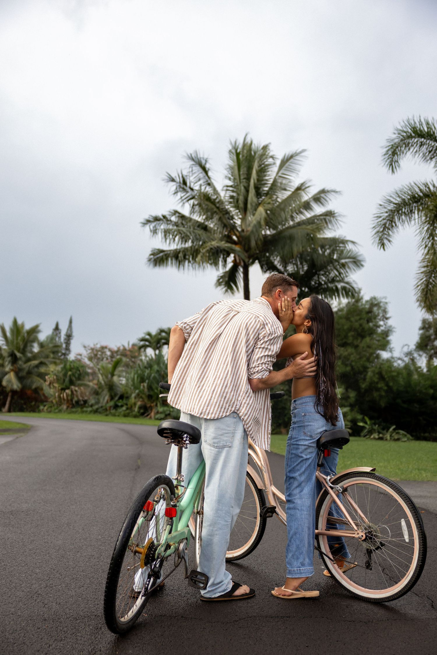 A couple shares a romantic kiss beside vintage bicycles on a palm-lined path.