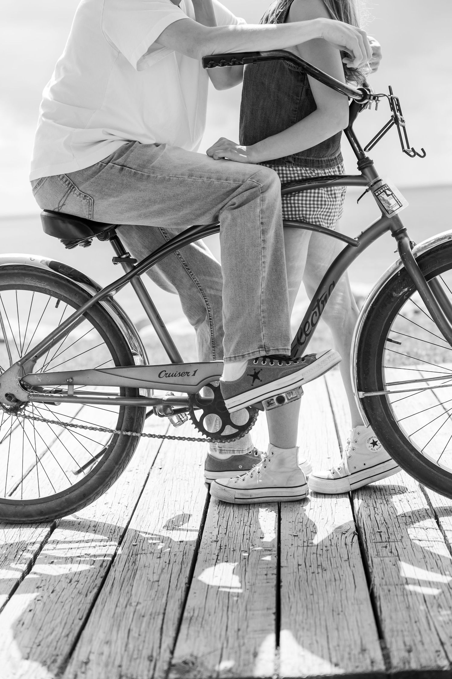 Black and white image of bicycle wheels and feet on a wooden boardwalk.