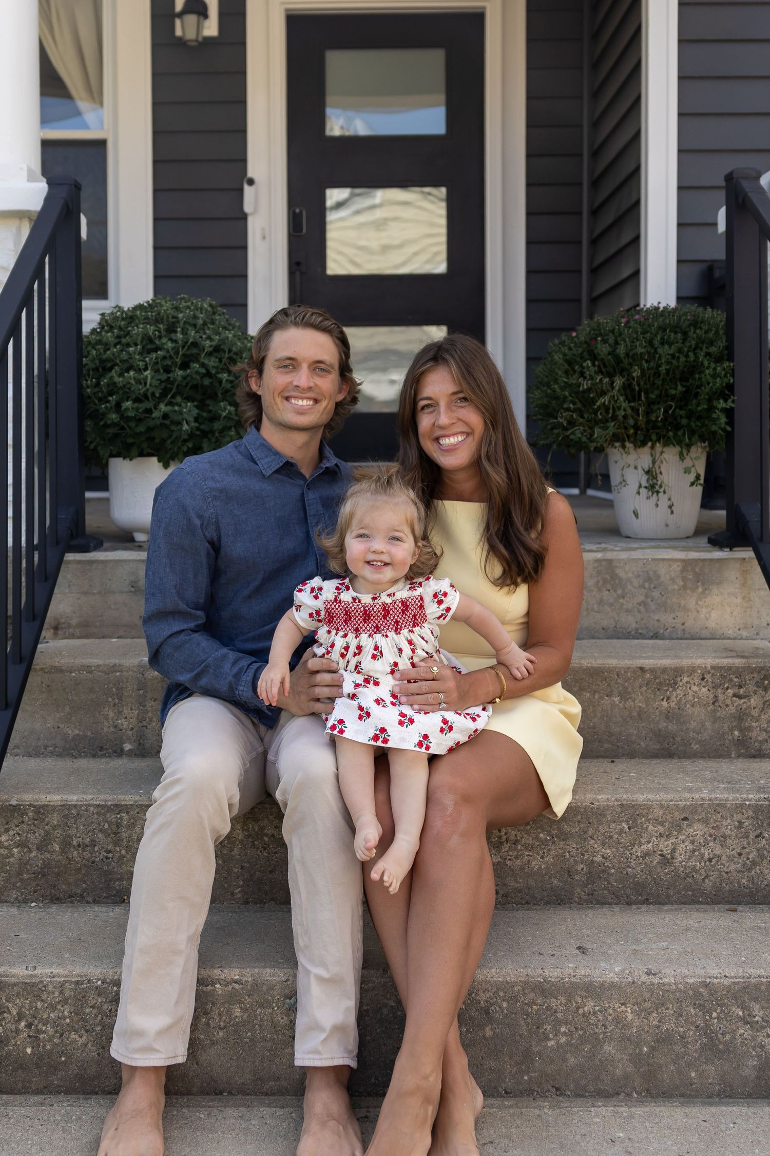 Family sits together on front porch steps of house with black siding.
