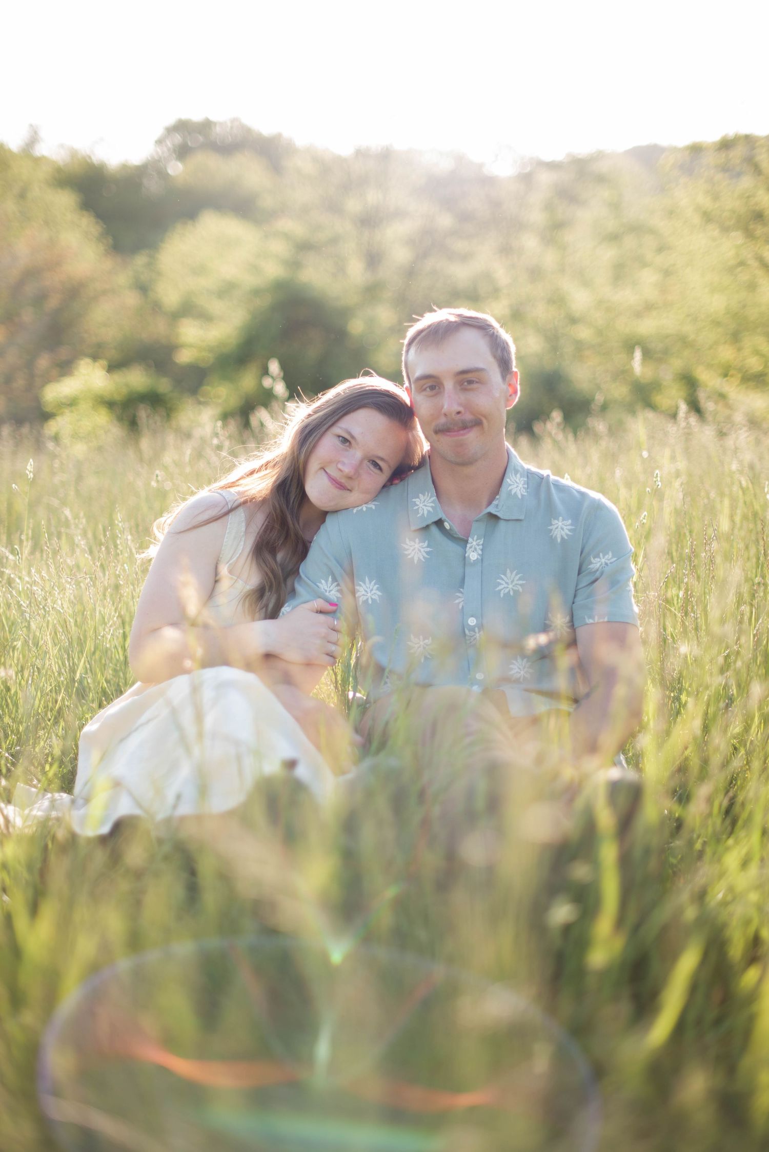 Couple sitting in field with mountain view looking at camera smiling