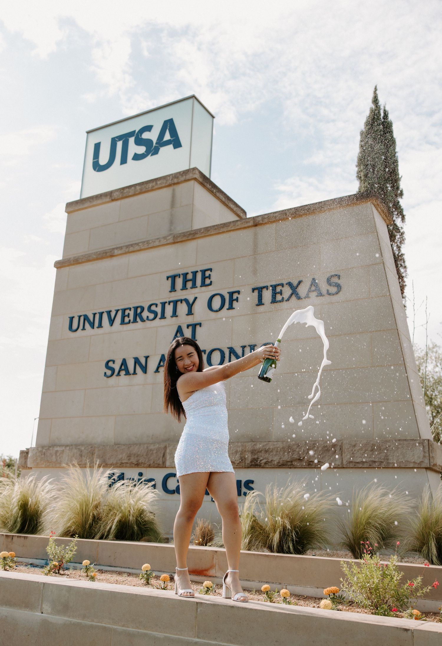 San Antonio Graduation Photography. Graduate celebrating in front of UTSA university entrance sign while splashing champagne.