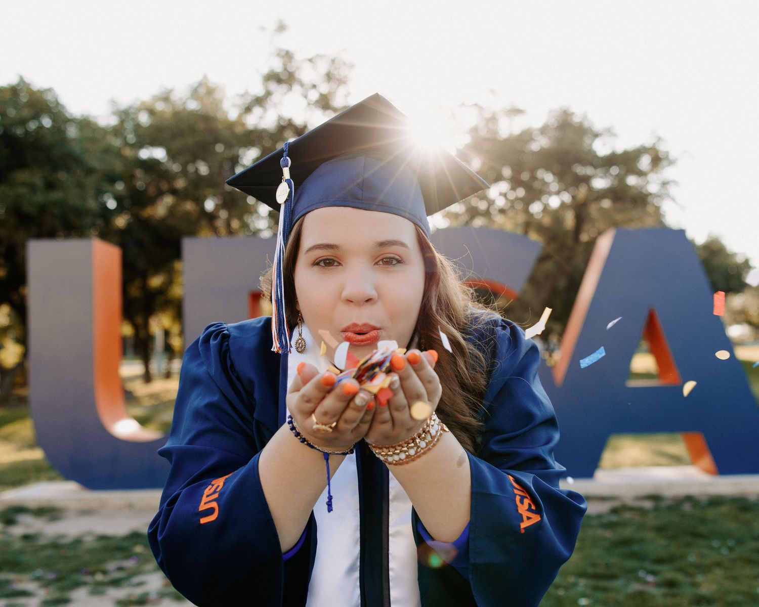San Antonio Graduation Photography. Graduate in blue cap and gown blowing glitter near UTSA letters during golden hour.
