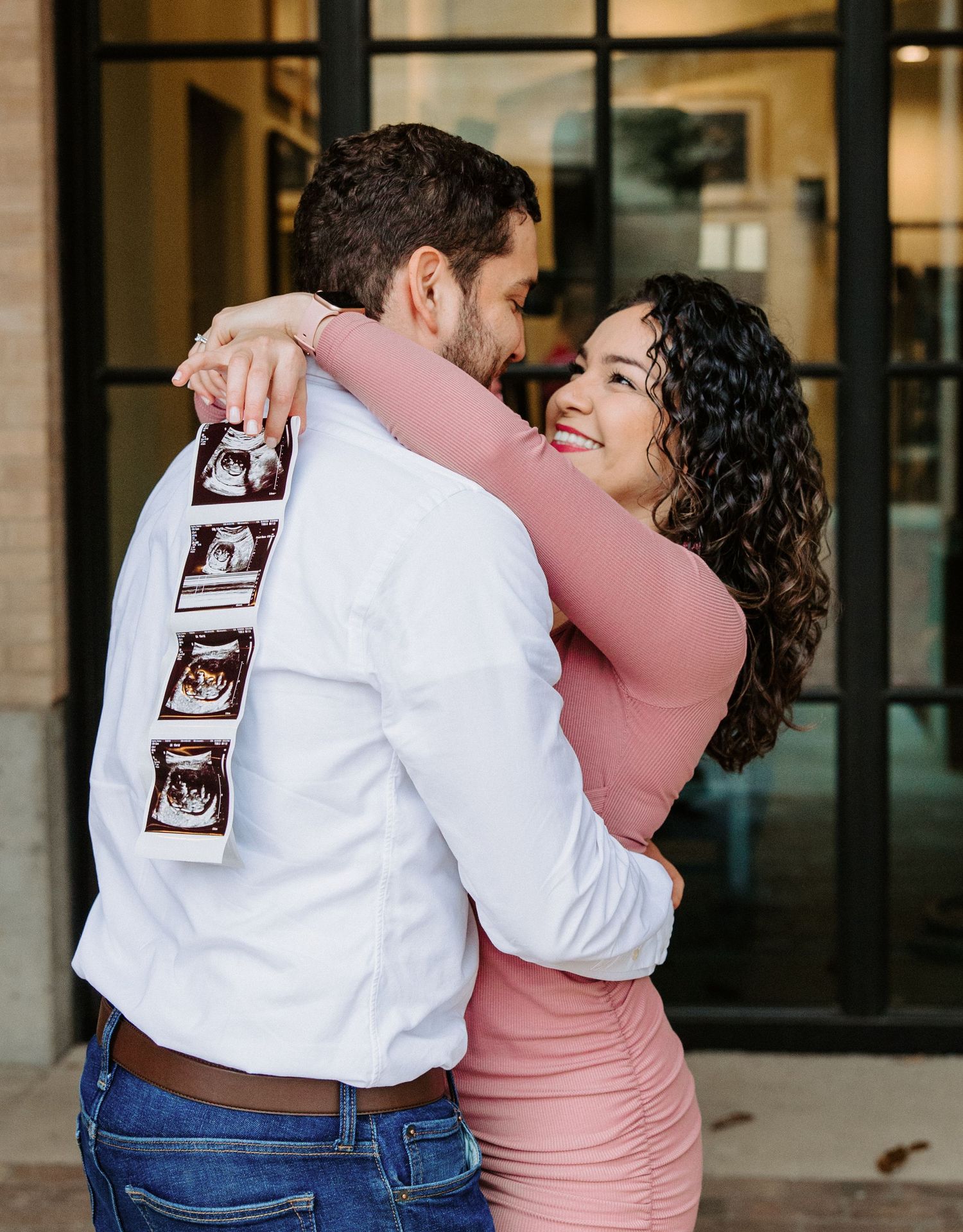 San Antonio Maternity Photography. Couple embracing and smiling while showing ultrasound photos on white shirt.