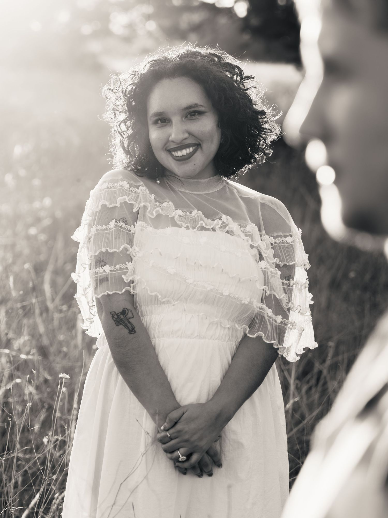 Black and white photo of a smiling bride in a lace white dress standing in a sunlit field.