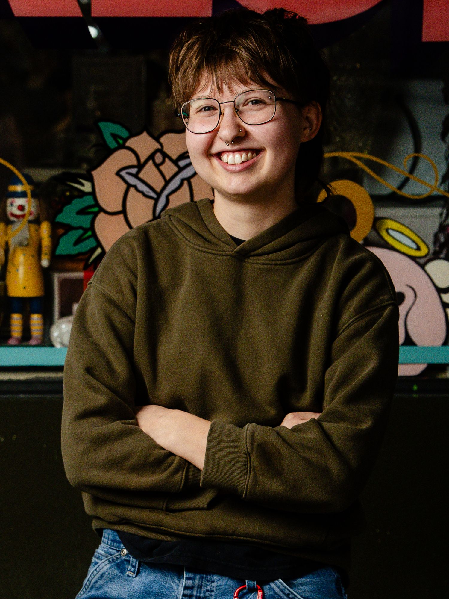 Person in olive green hoodie and jeans sits casually on wooden surface in front of graffiti wall.