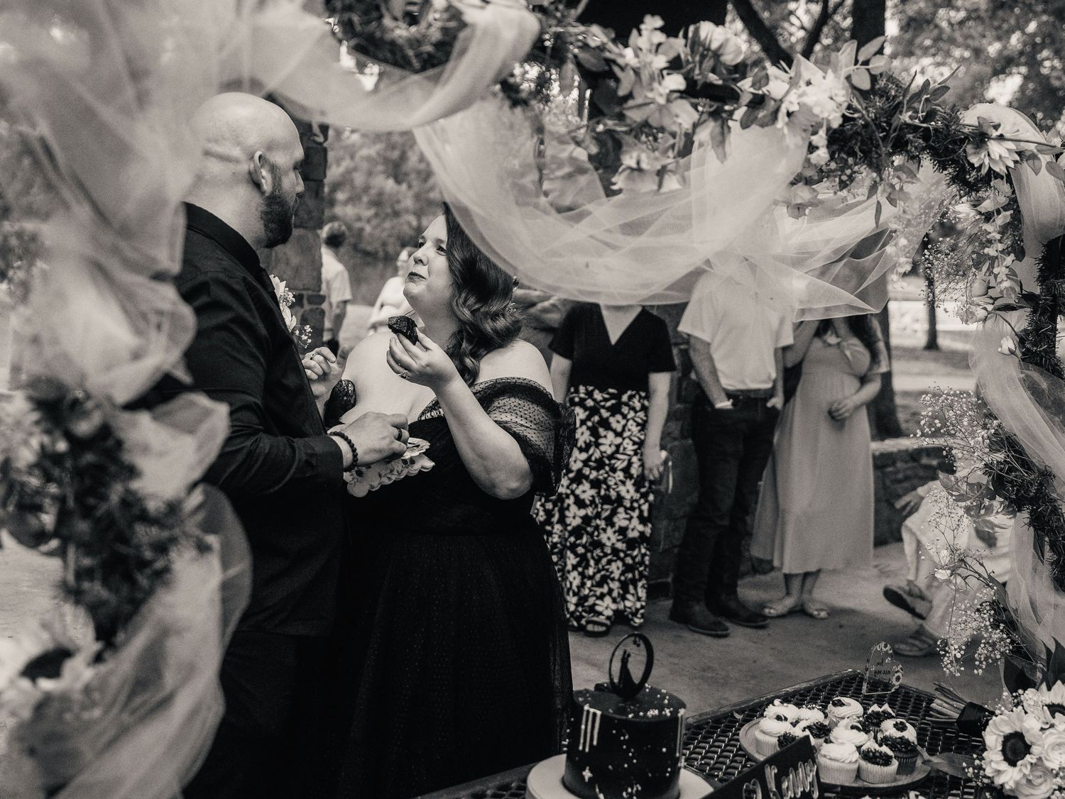 Black and white wedding photo of guests laughing as a veil floats through the air during an outdoor ceremony.