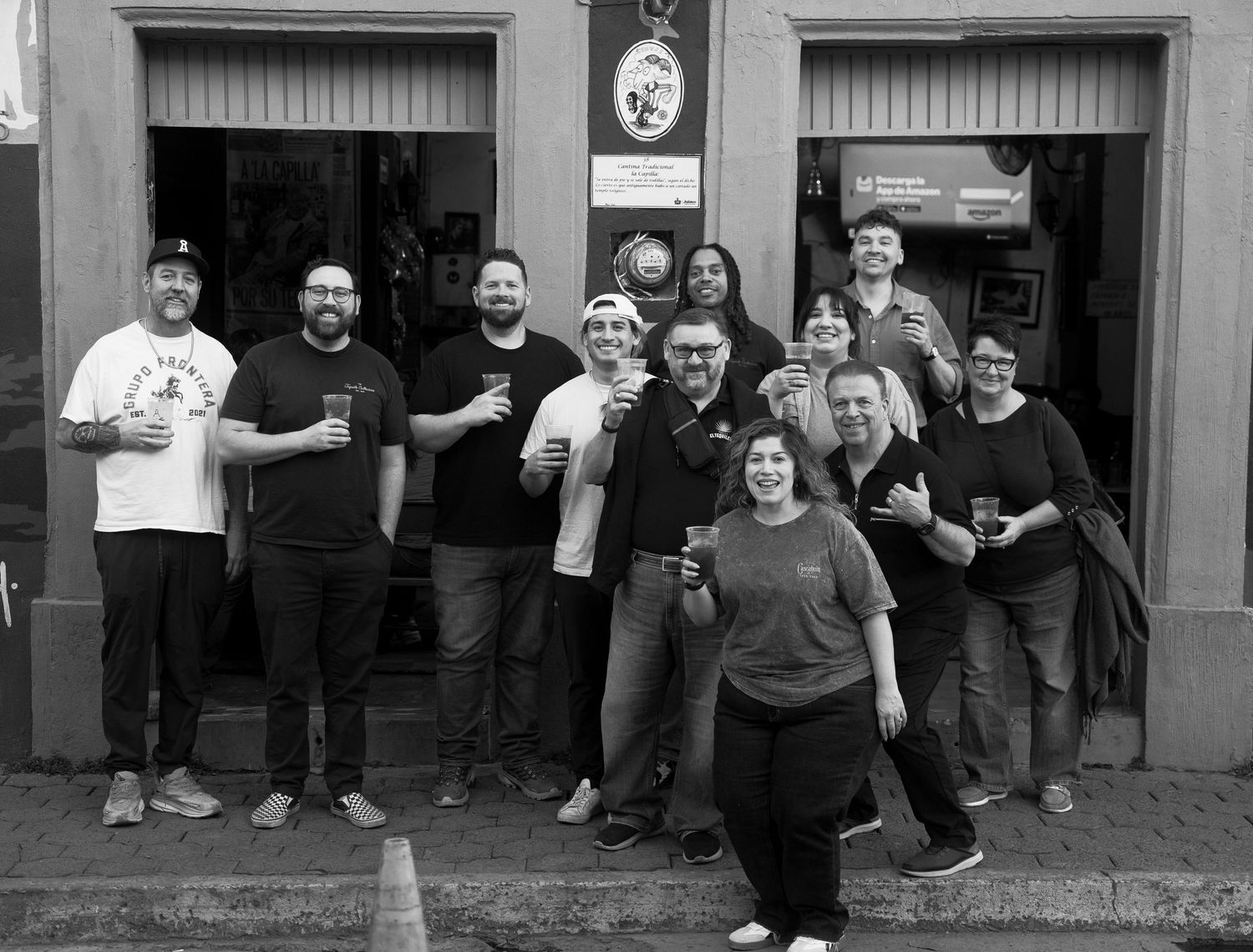 Black and white photo of a large group of friends posing together outside a bar, holding drinks and smiling.