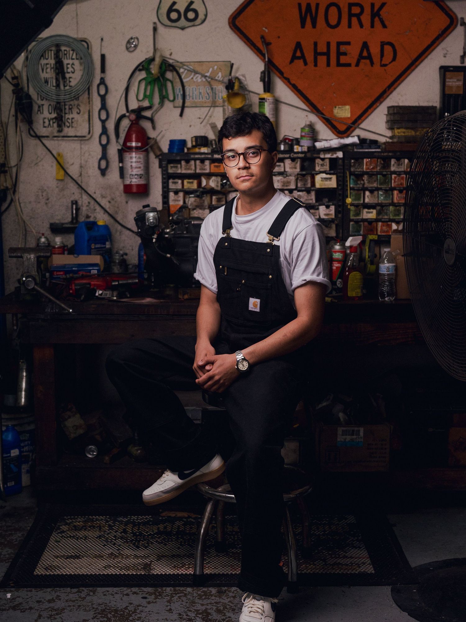 Young person in overalls sits in a vintage garage workshop with a Work Ahead sign and Route 66 memorabilia.