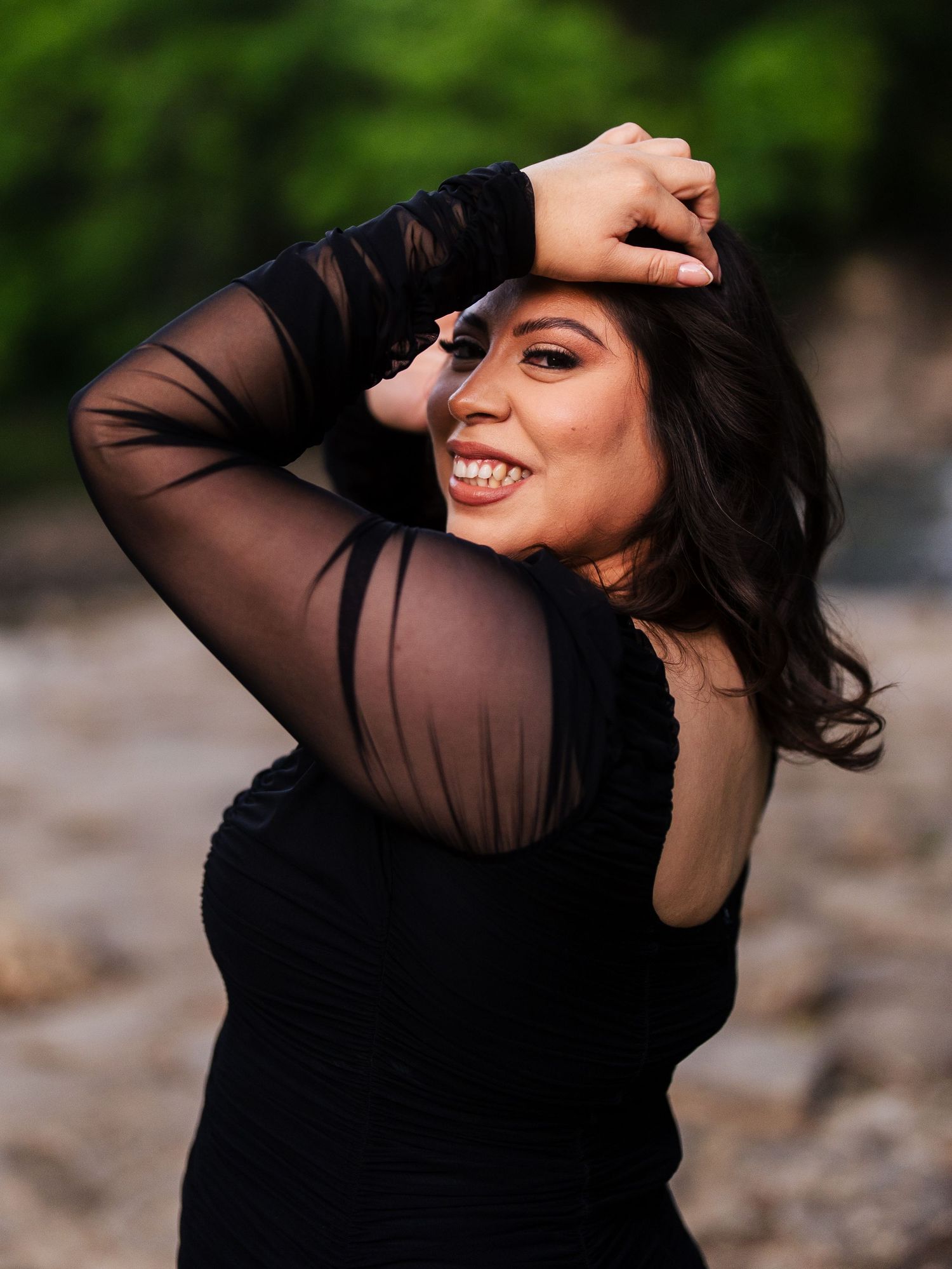 Person in sheer black long sleeve dress poses outdoors while smiling and running hand through dark wavy hair.