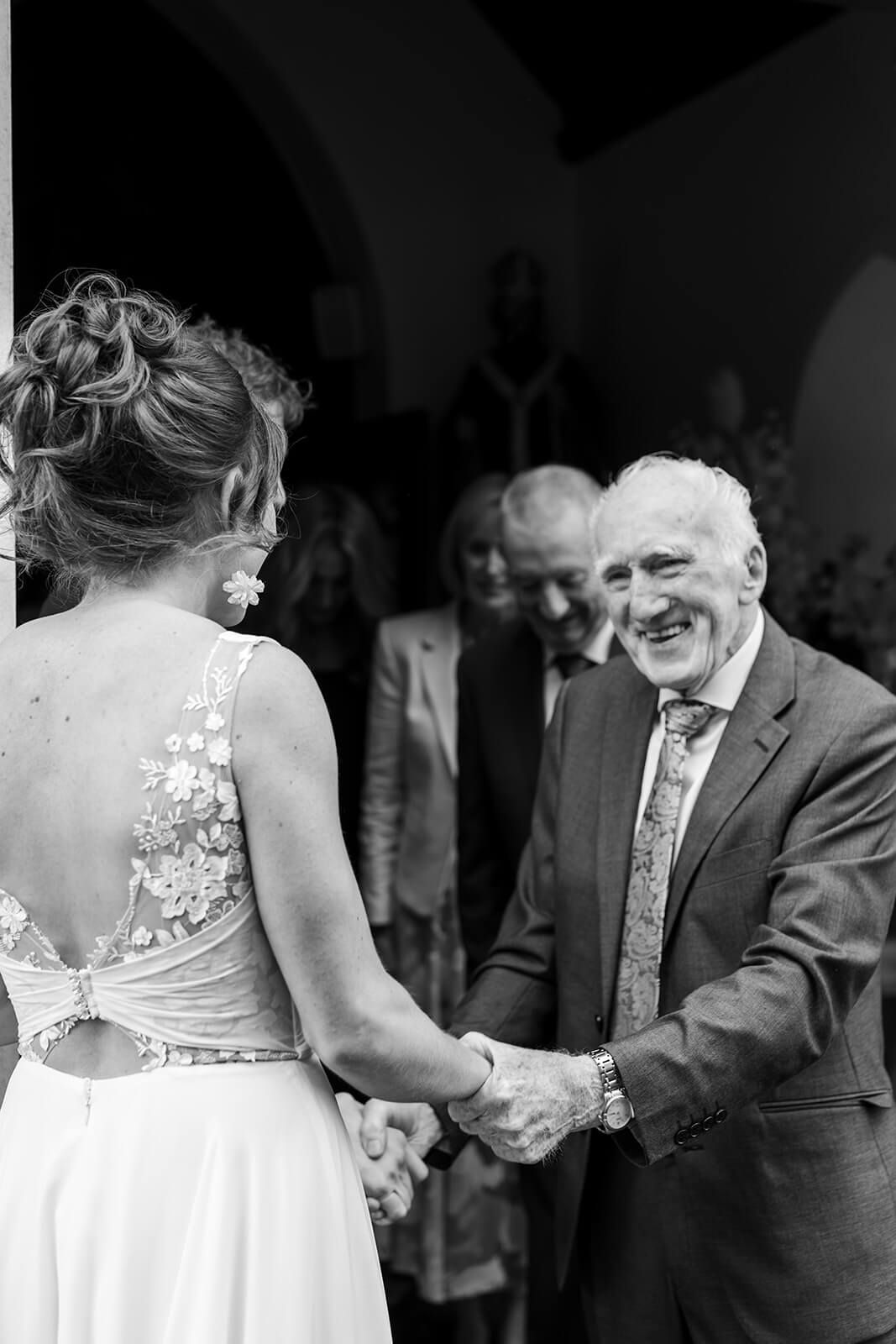 A touching black and white wedding moment showing a bride in a lace dress greeting an elderly guest with a handshake.