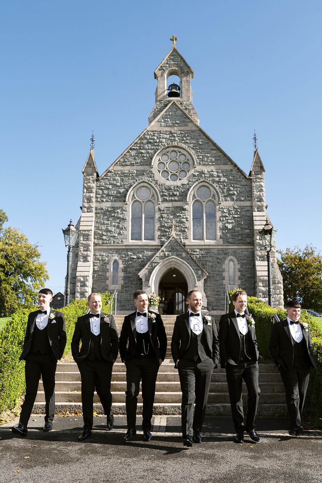 A group wearing black tuxedos stands on stone steps in front of a historic stone church with Gothic architecture.