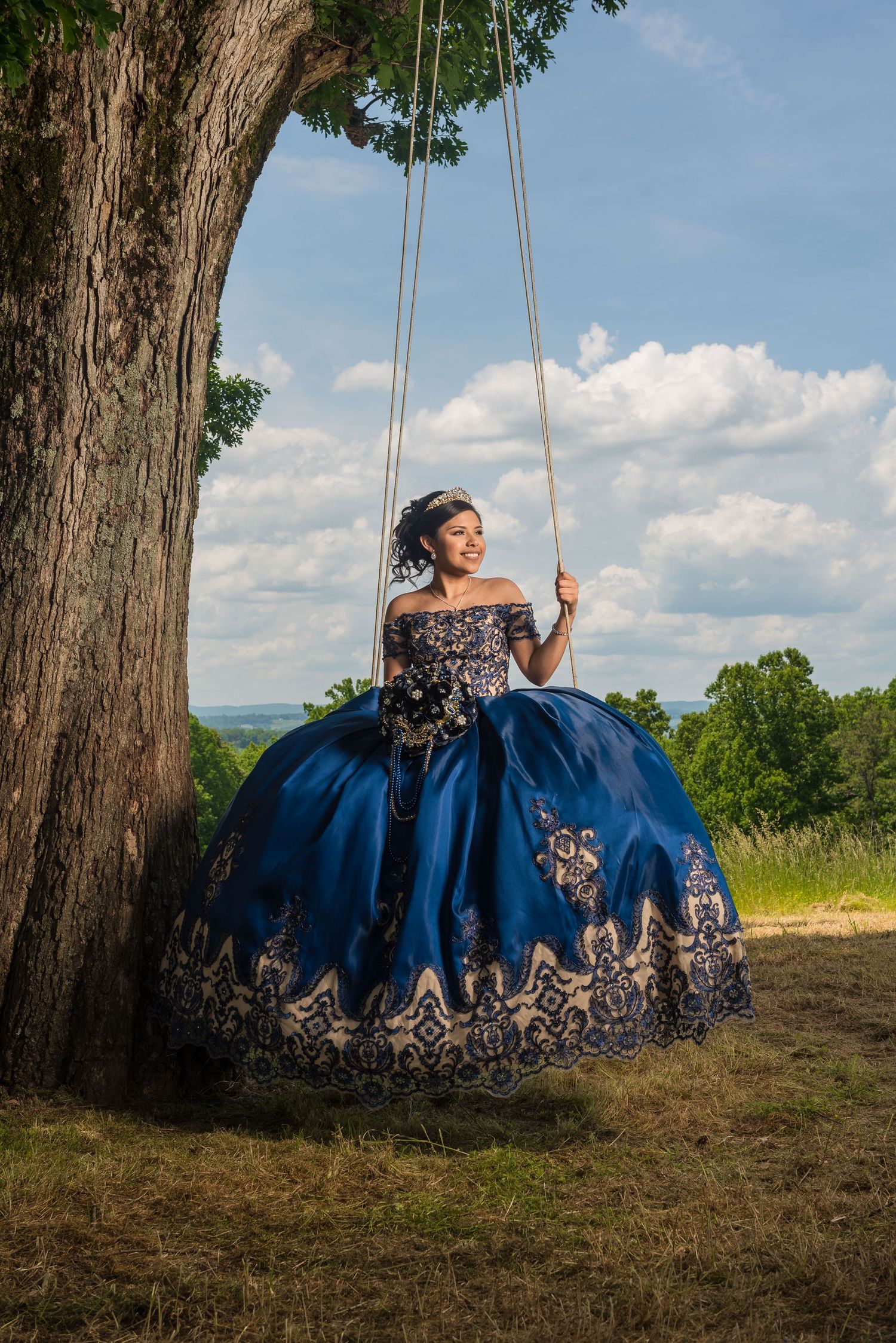 A person in an elegant navy blue quinceañera dress poses gracefully on a tree swing against a scenic outdoor backdrop.