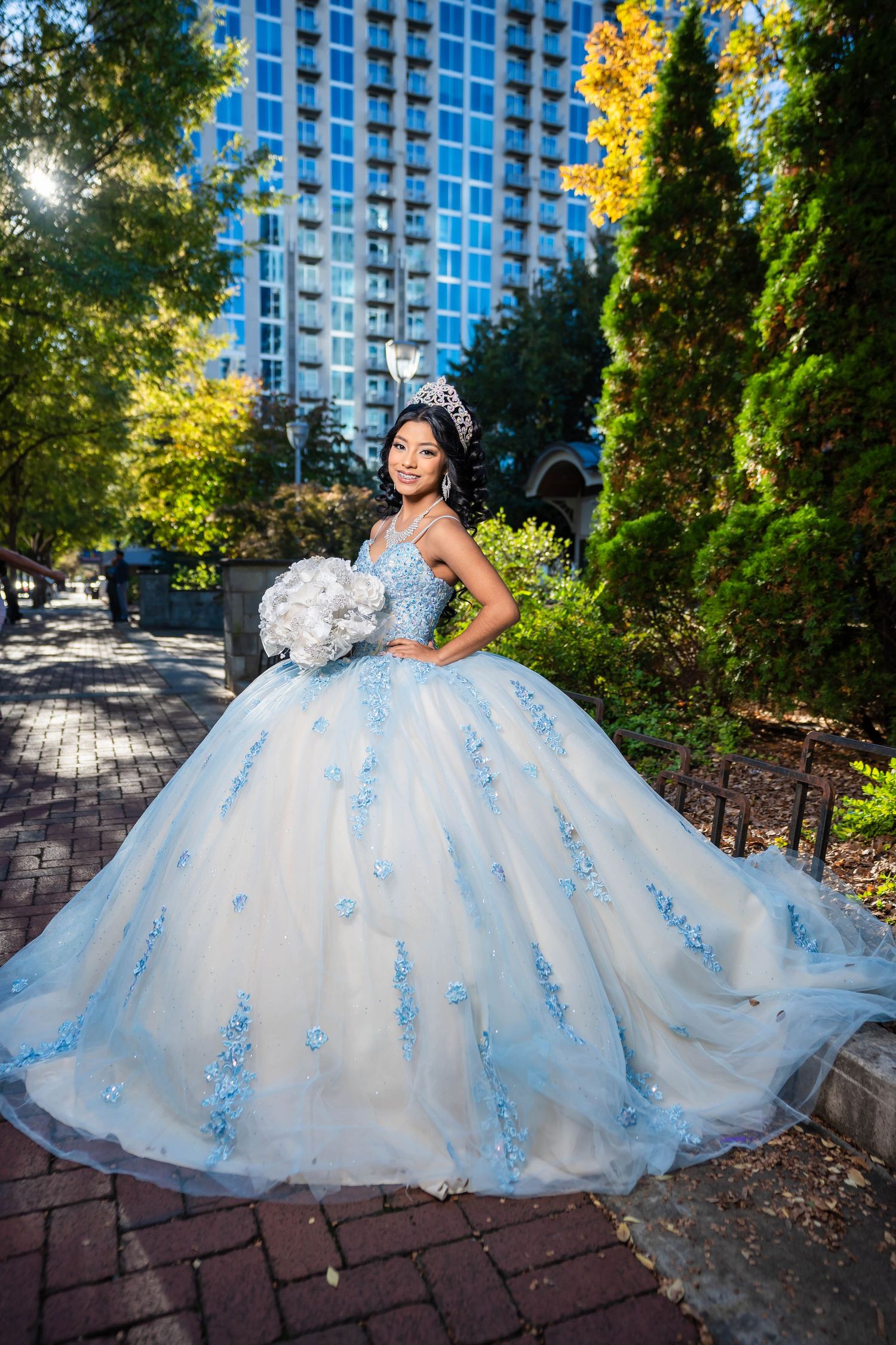 A stunning white and blue floral ball gown photographed in a garden setting with brick pathways and tall buildings in background.