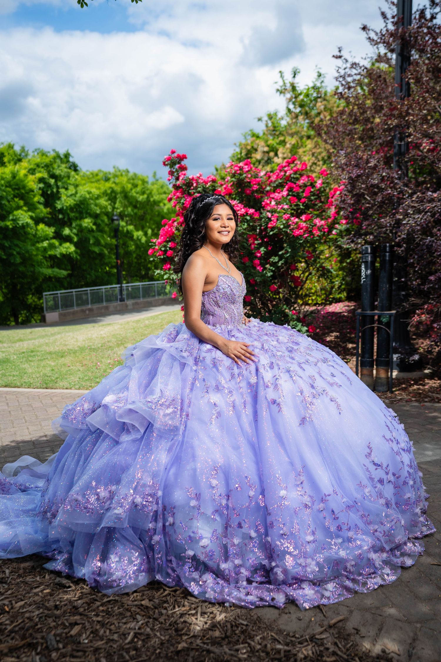 A lavender ballgown with floral appliques sparkles in an outdoor garden setting with blooming pink roses and greenery.
