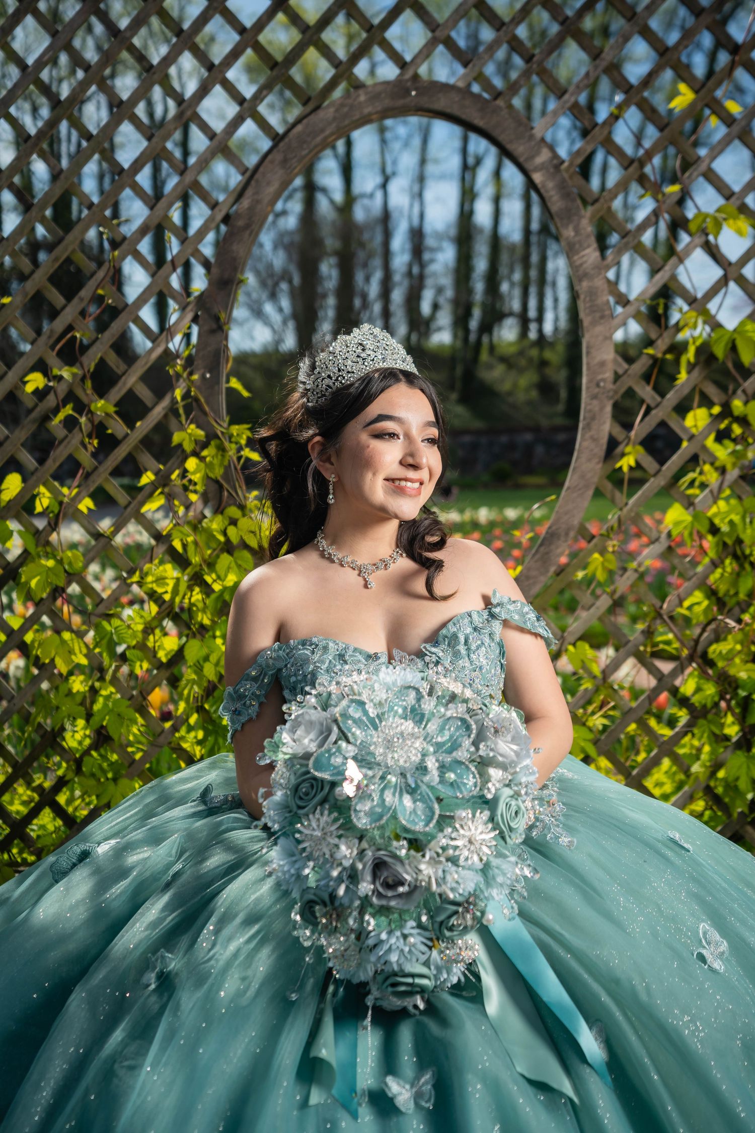 A person in a sparkling turquoise quinceañera dress poses gracefully against a garden lattice backdrop.