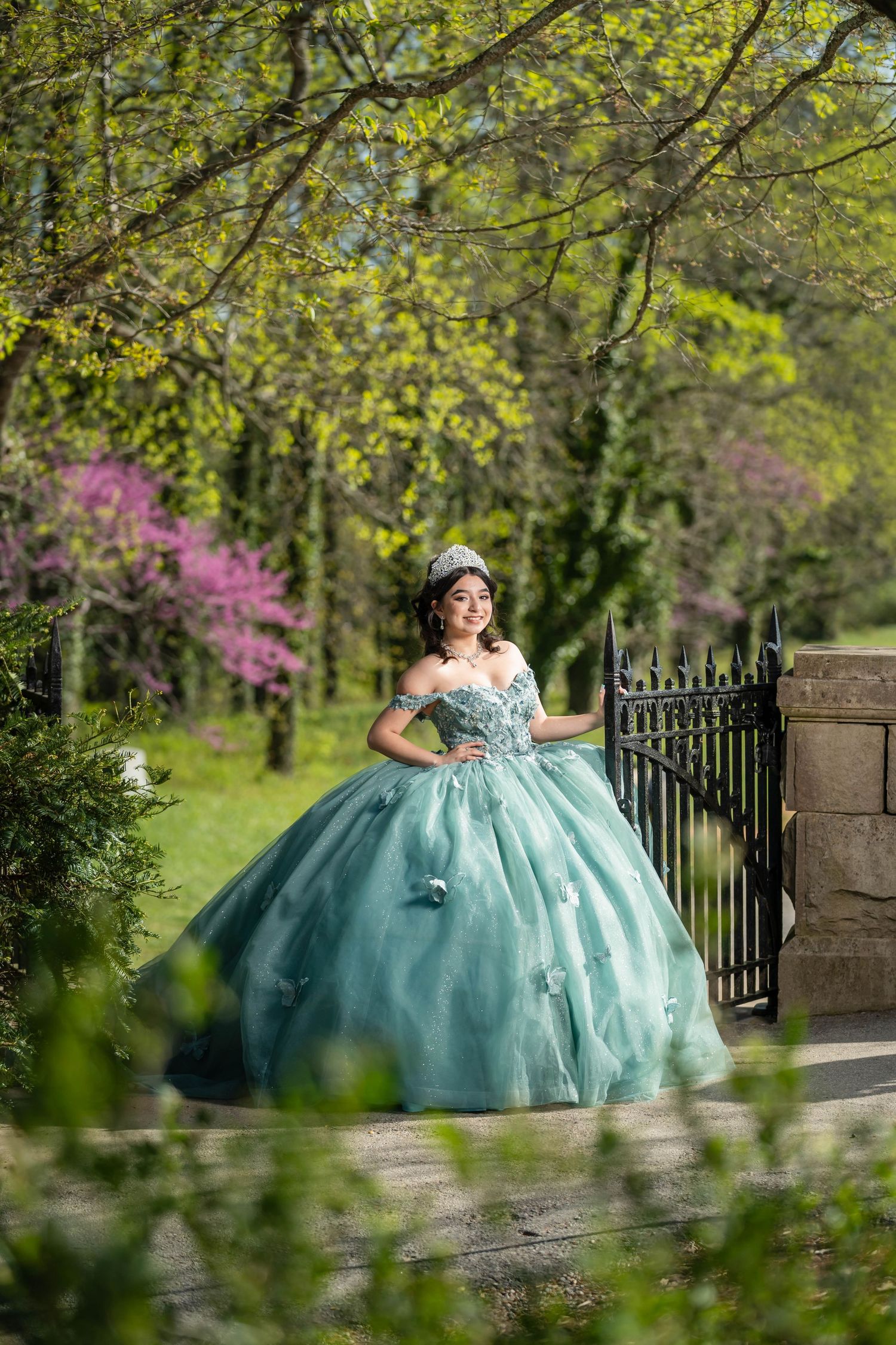 A quinceañera in a turquoise ball gown poses elegantly in a spring garden with blooming trees and wrought iron gates.