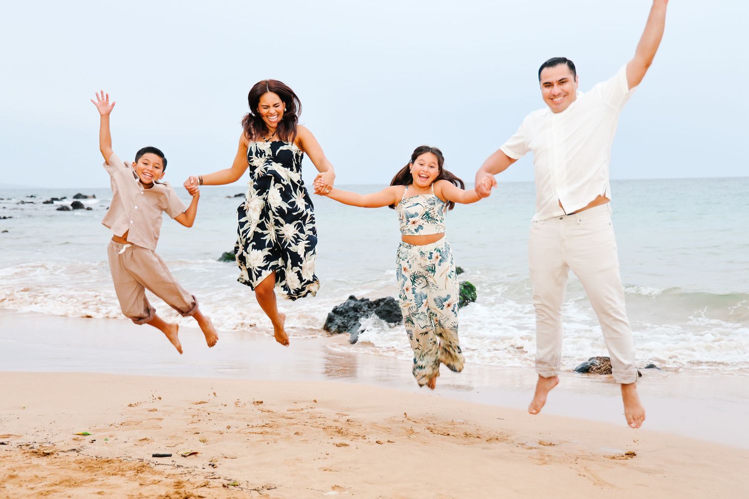 Family wearing casual outfits jumps joyfully together on a sunny beach with ocean waves in the background.
