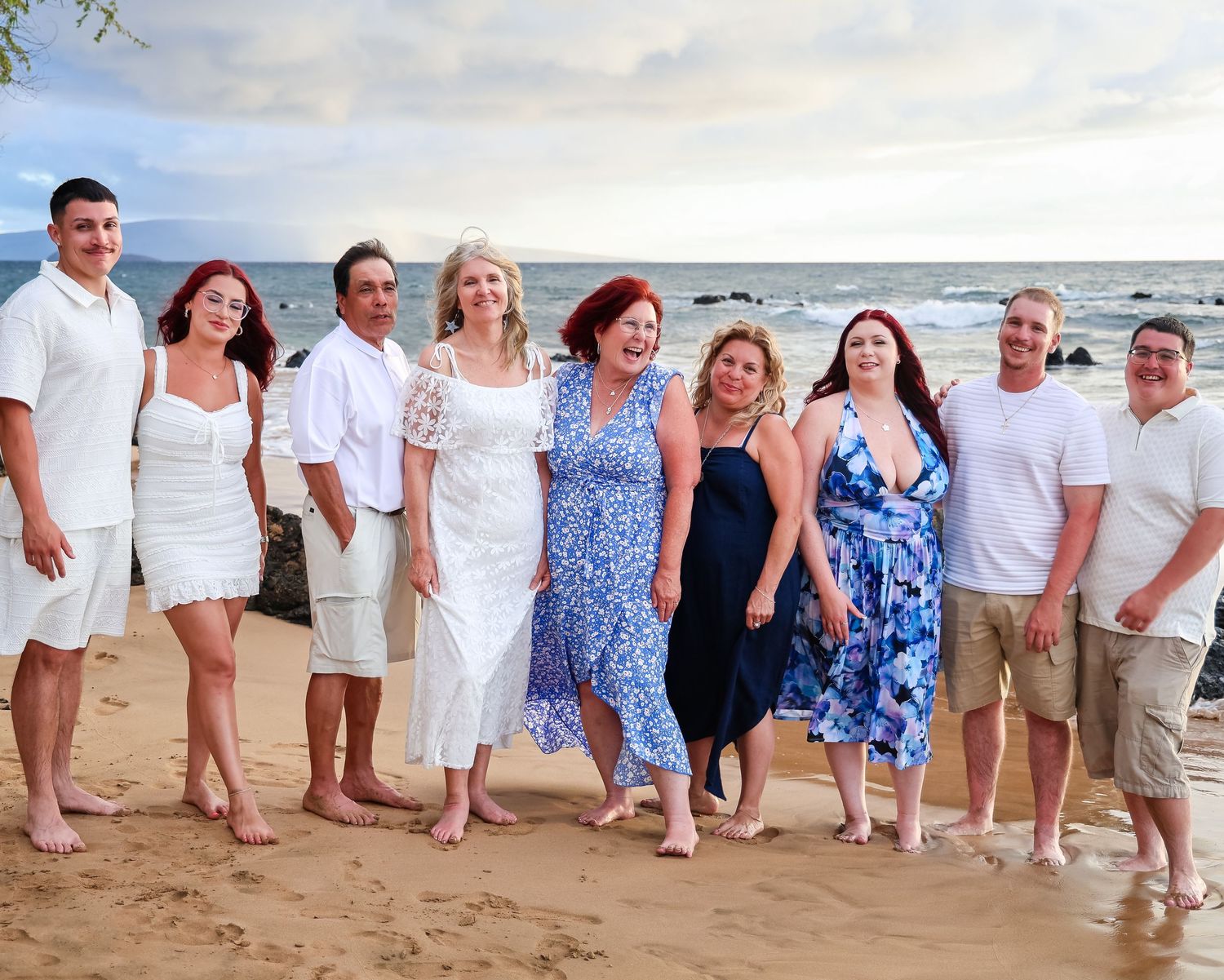 A group of people in coordinated blue and white outfits pose together on a sandy beach at sunset.