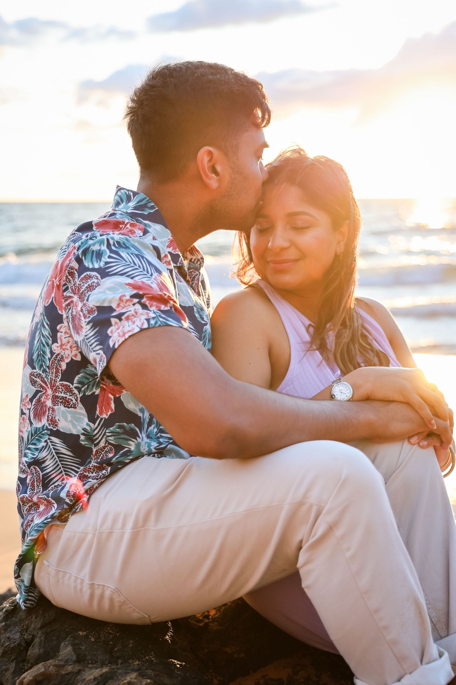 Couple sharing a romantic moment at sunset on a beach, man kissing woman on cheek while sitting on rocks.