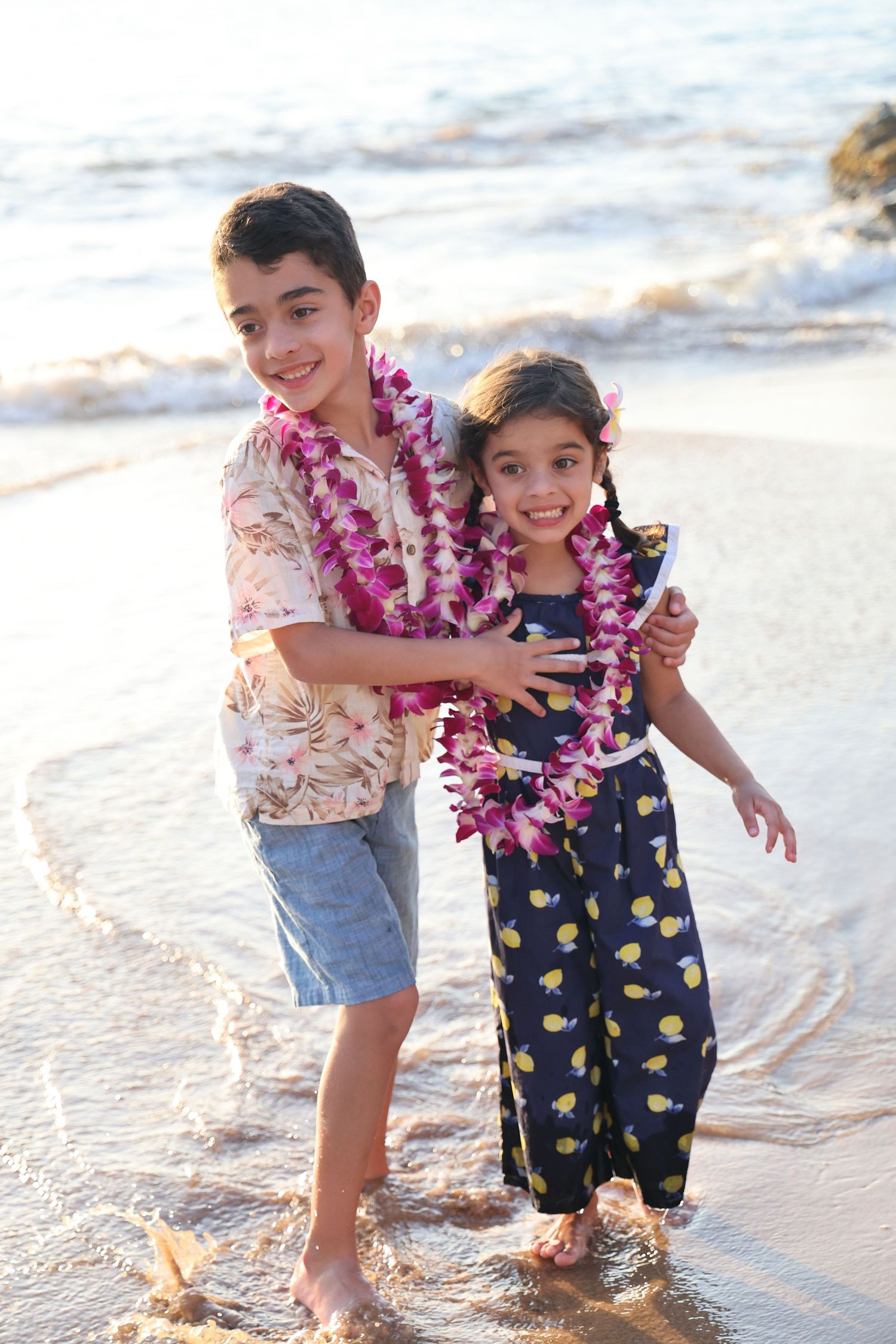 Two children wearing leis and casual clothes play together at the beach during sunset.