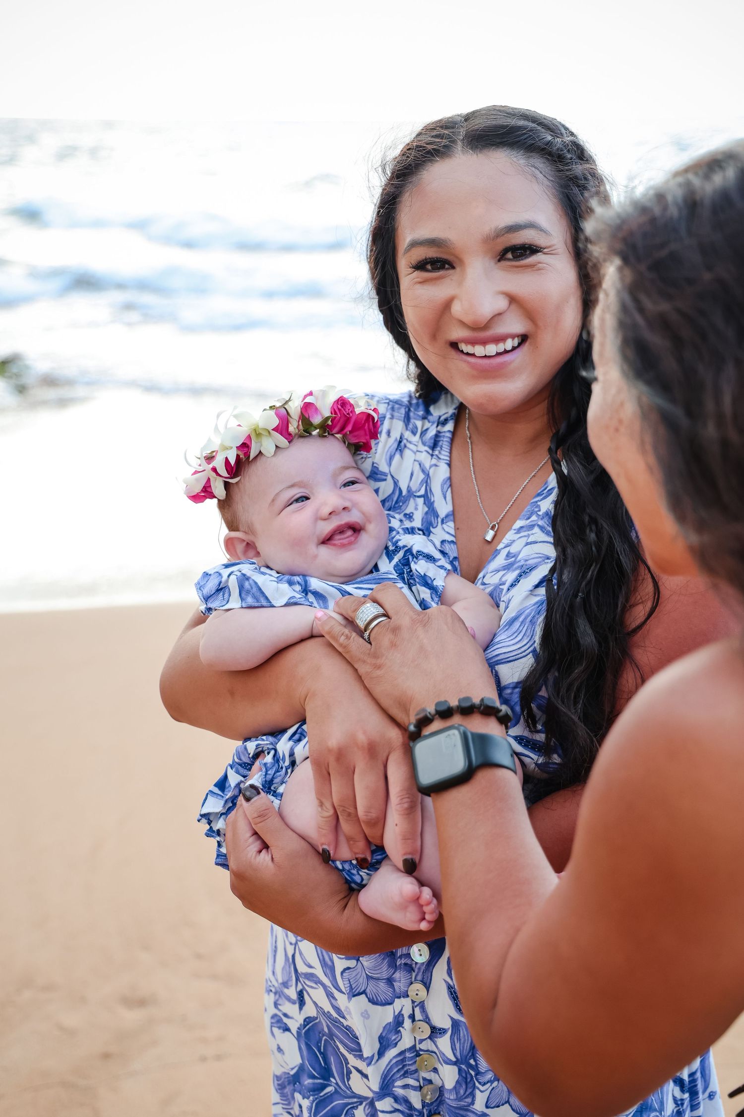 Smiling woman in blue floral dress holds a baby with flower crown on sandy beach.