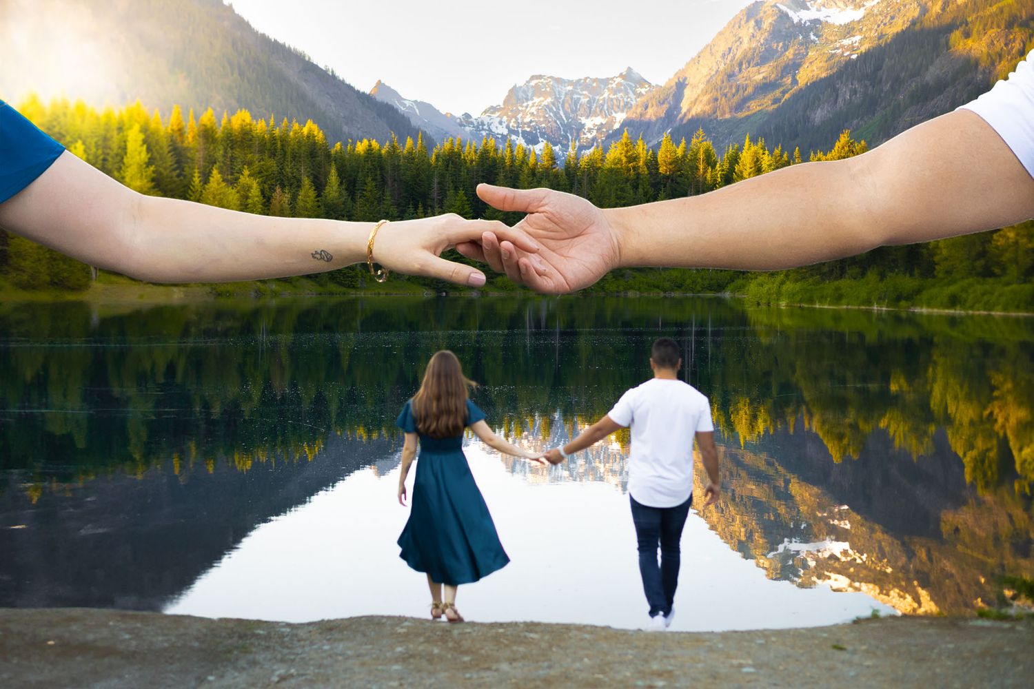 A young couple holding hands walk toward Gold Creek Pond with a mountainous backdrop during an engagement session.