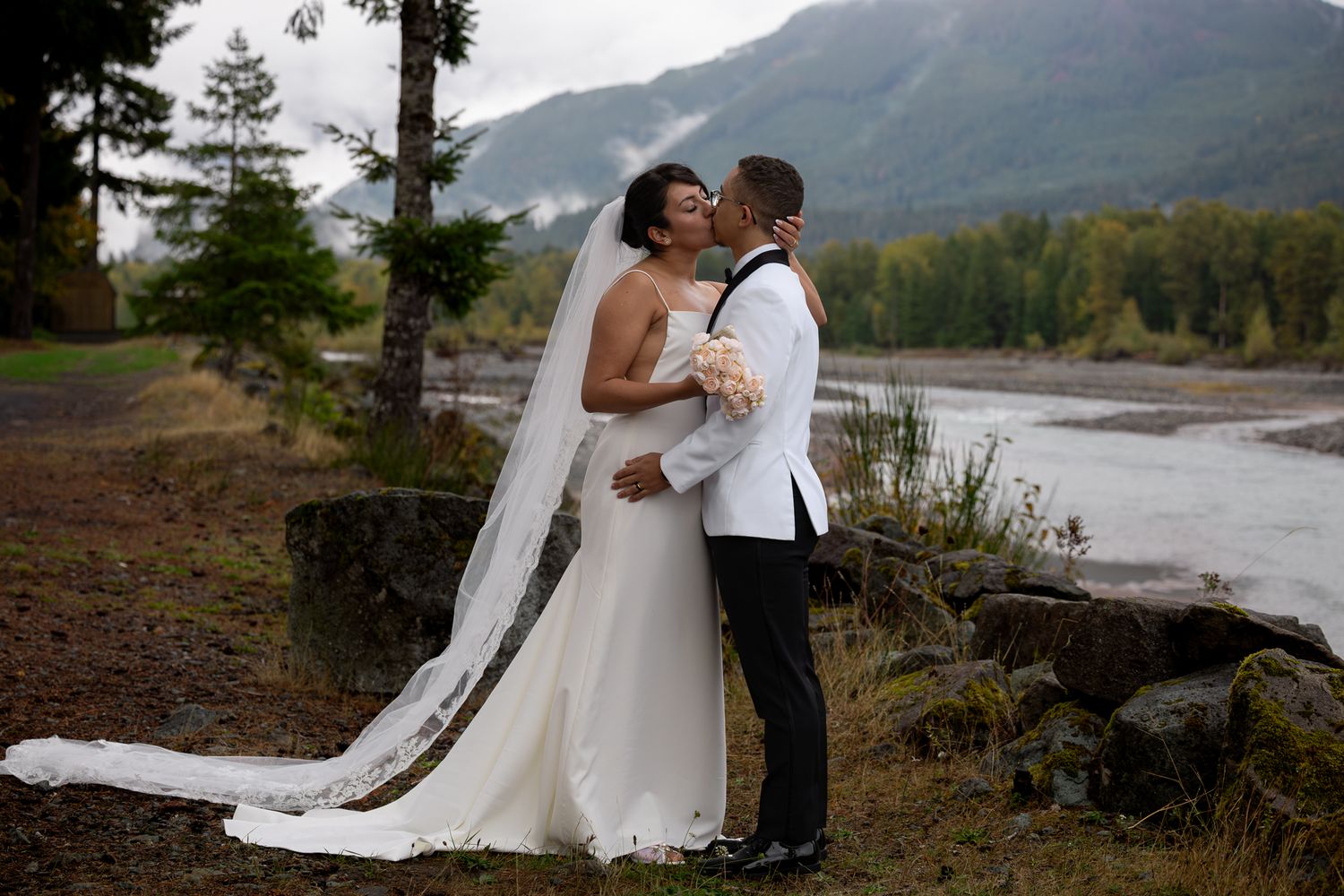 Bride and groom's first kiss with the backdrop of the turquoise tinted nisqually river and a moody mountainous landscape blanketed with fog.