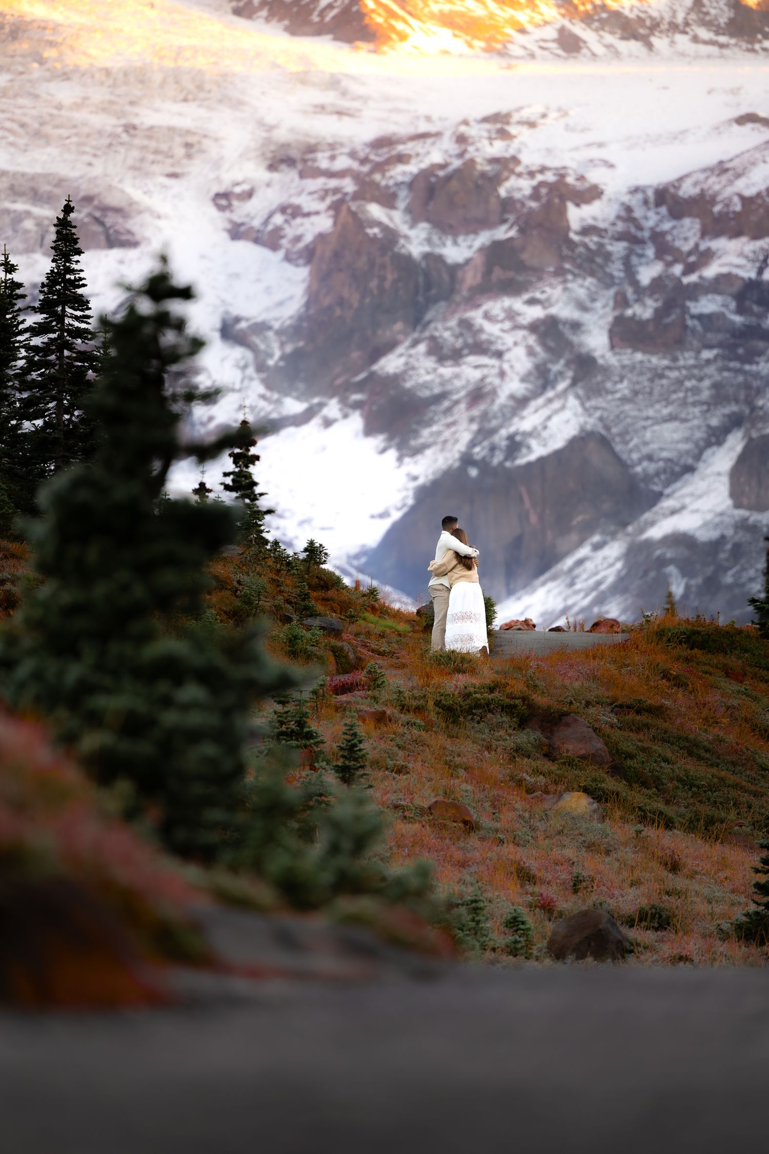 Dramatic backdrop of Mount Rainier, glowing at sunrise, with fall foliage. A couple embrace on a trail for their engagement session.