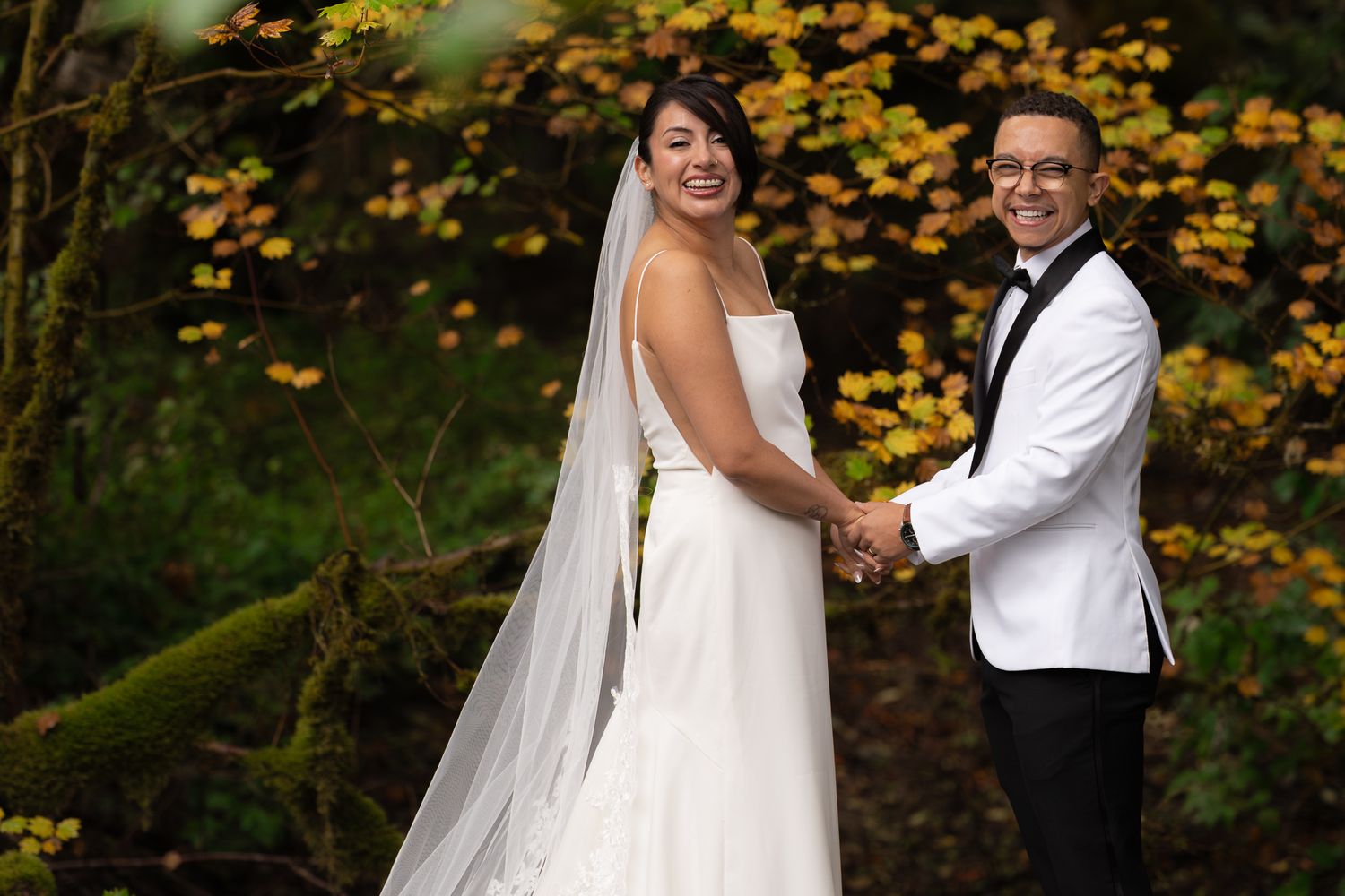 Surrounded by fall foliage, a couple smile at their wedding photographer after doing first looks on their wedding day near Mount Rainier.