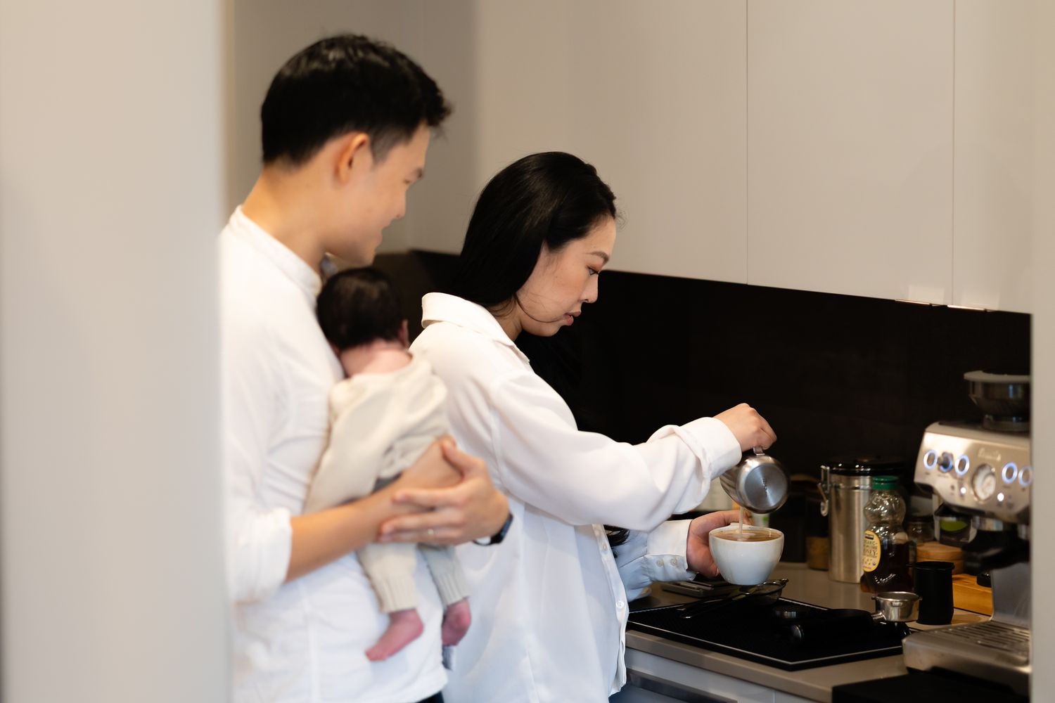 New mom Amy makes a latte on her Breville while dad holds their newborn during an in-home lifestyle newborn session near Seattle.