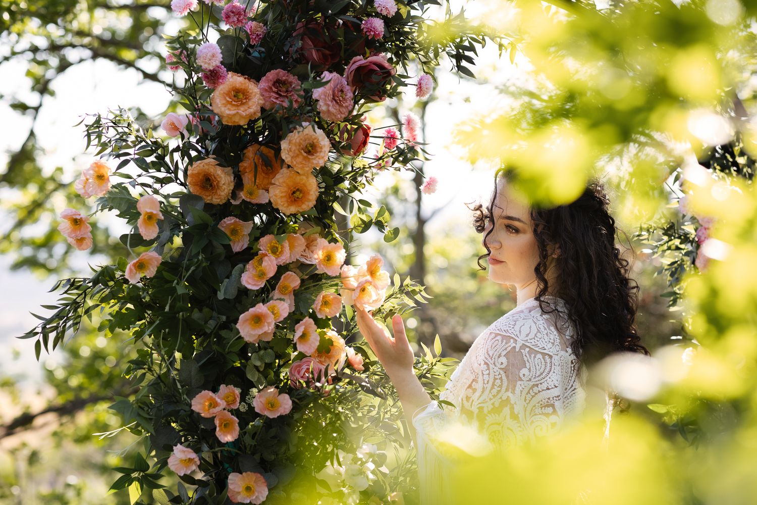 A boho bride reaches for a colorful flower arch. Wind blows her curls over her face and she is framed by sun-dappled trees.